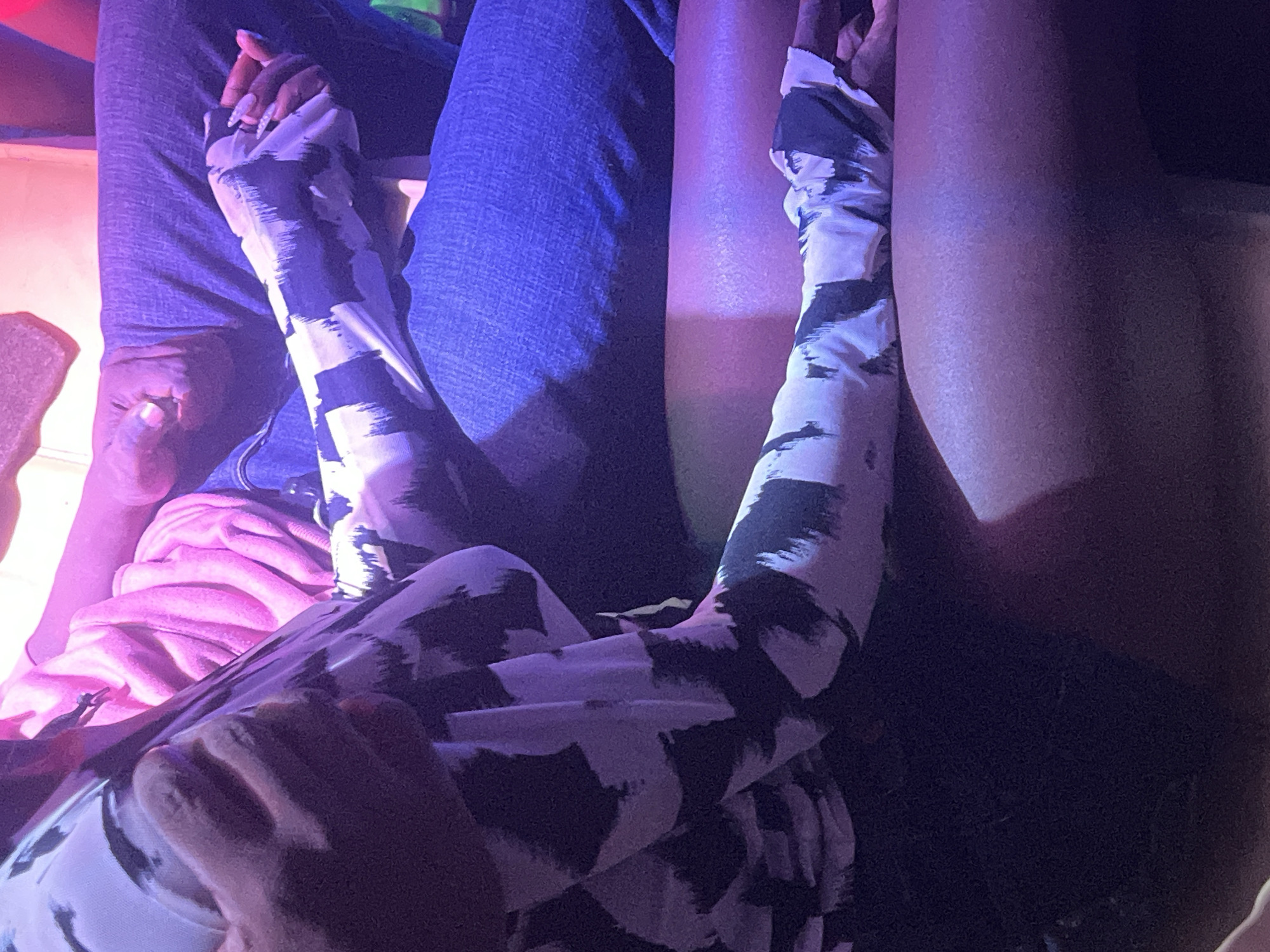This is a close-up, low-angle shot of two people sitting next to each other, likely in a dimly lit indoor space with purple ambient lighting. The person in the foreground, on the left, is wearing a black and white patterned long-sleeved shirt. Their left arm is visible, resting on their lap. The right arm of this person is bent and their hand is clasped with the hand of the person next to them. The person on the right is wearing blue jeans. Their left arm is also visible, resting on their lap and is partially covered by the sleeve of the person on the left. The right arm of the person on the right is extended and their hand is interlaced with the hand of the person on the left. A pink garment, possibly a sweater or hoodie, is visible behind the person on the left, suggesting they are seated in a row of seats or a booth. The overall scene is intimate and suggestive of a shared moment between the two individuals. The image does not provide any information to indicate its location in Gura Top, Nigeria.