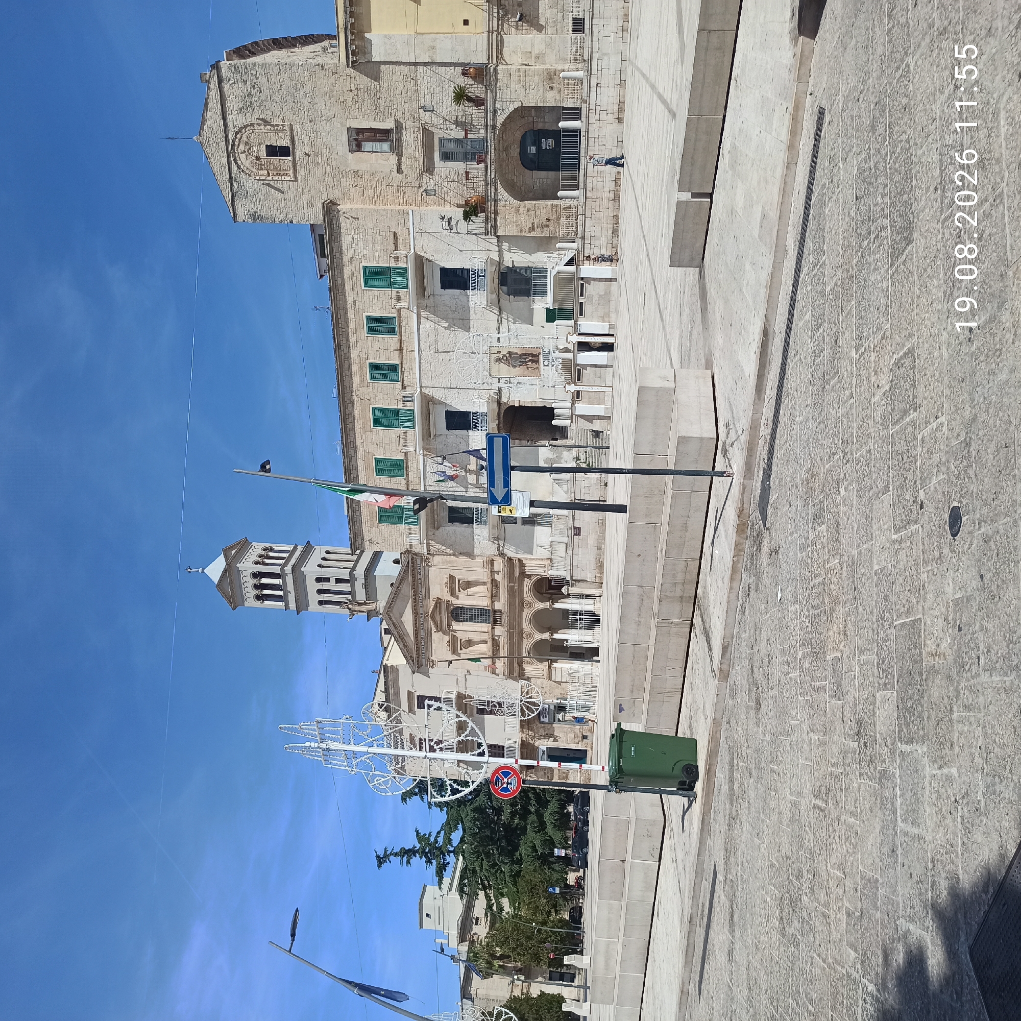 This is a photograph of a town square in Ruvo di Puglia, Italy, on a bright, sunny day. In the background, a large, light-colored stone building with a bell tower dominates the scene. The building has multiple windows with green shutters and decorative architectural elements. In front of it, a street or plaza is paved with cobblestones.
On the right side of the square, there are white, decorative, lattice-like structures, possibly for holiday decorations, and a green trash bin. A lamppost stands to the left of the trash bin, and further left, another lamppost is visible. A sign with a red circle and a diagonal line, indicating a prohibition, is mounted on a pole near the lamppost.
A few people are present in the square. Two individuals are walking in the middle ground, and another person is walking in the far right background. The sky is a clear blue with a few thin clouds. The overall scene depicts a public space in an Italian town, likely a historically significant area given the architecture. A date stamp in the lower right corner reads "19.08.2026 11:55".