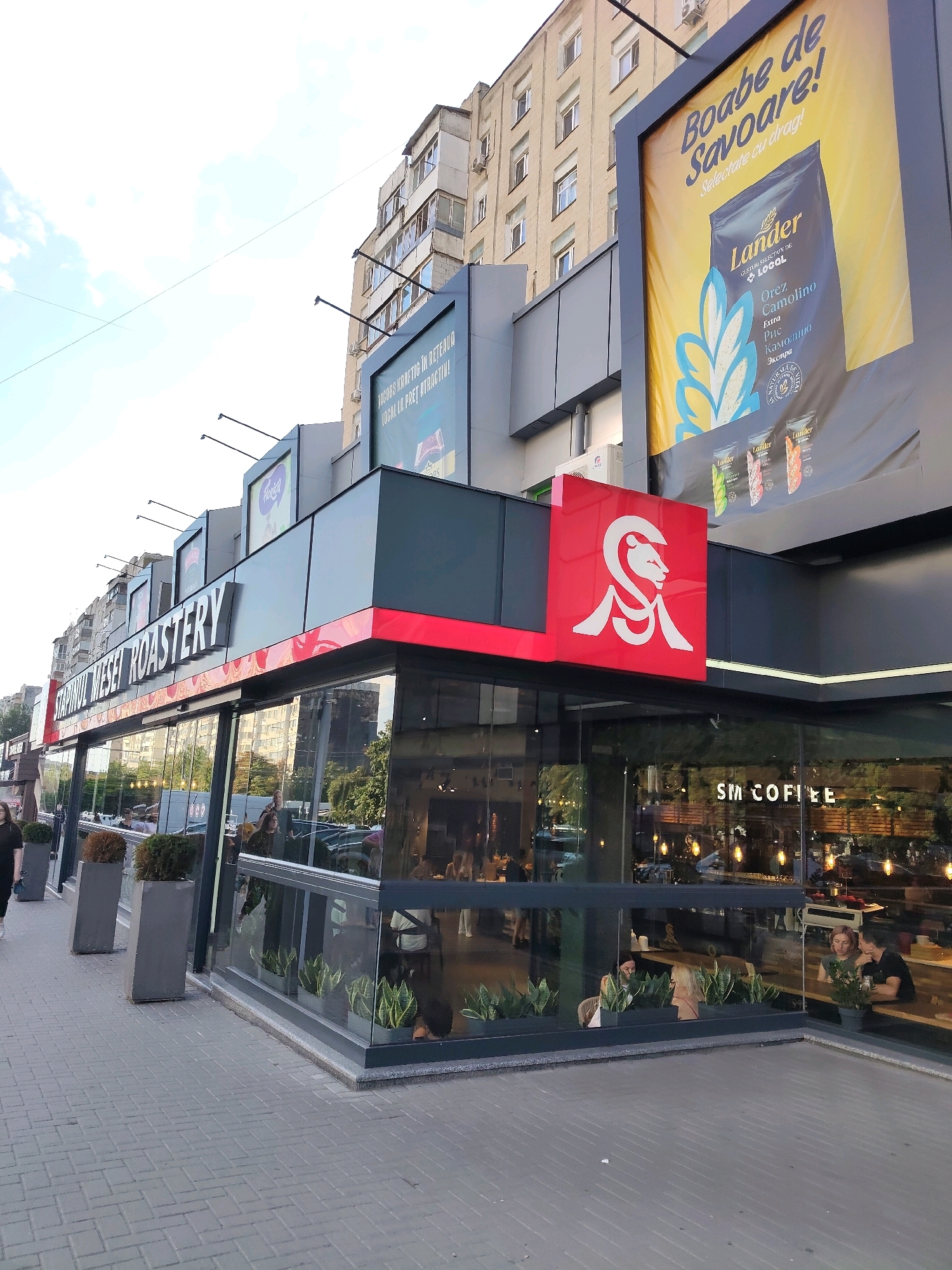 The image depicts a street-level view of a commercial establishment, possibly a coffee shop or roastery, in an urban setting. The building features a modern facade with large glass windows and prominent signage. A large, illuminated red sign with a stylized white lion emblem is mounted on the corner of the building. Above the entrance, a large white sign reads "STAPINULUI MESEI ROASTERY." A large billboard is mounted above the building's roofline, advertising "Boabe de Savoare!" with an image of a rice bag labeled "Lander Orez Camolino." The street in front of the building is paved with gray rectangular bricks. Planters with greenery are visible outside the glass windows. Inside the establishment, several people are seated at tables, and reflections in the glass show interior lighting and decor, including a sign that reads "SM COFFEE." A building with multiple stories and visible windows rises in the background. The sky is visible with some clouds.