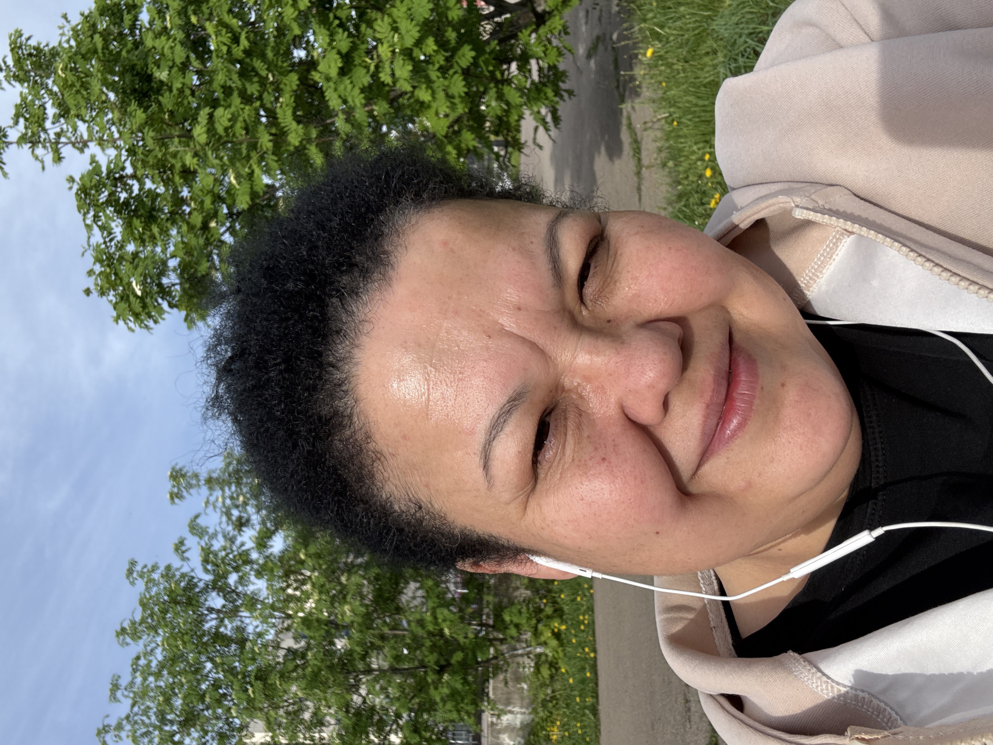 A close-up, vertically oriented photograph shows a woman outdoors in Unknown. She is wearing a light pink hooded sweatshirt over a black shirt. White earbuds are visible, with one inserted in her left ear. The background includes green trees and a sidewalk. The sky is visible in the upper left corner, indicating daylight.