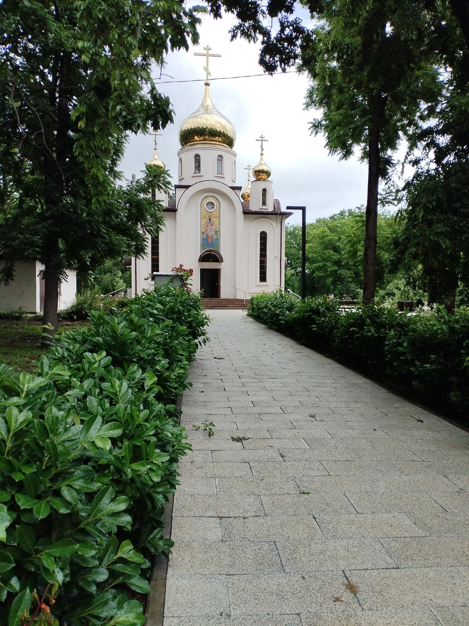 In Vinogradny, Russia, a paved walkway leads towards a white Orthodox church. The church features golden domes topped with crosses and a mosaic above the arched entrance. Lush green bushes line the walkway on the left, and mature trees surround the scene, creating a dappled light effect. The sky is overcast. No people are visible.