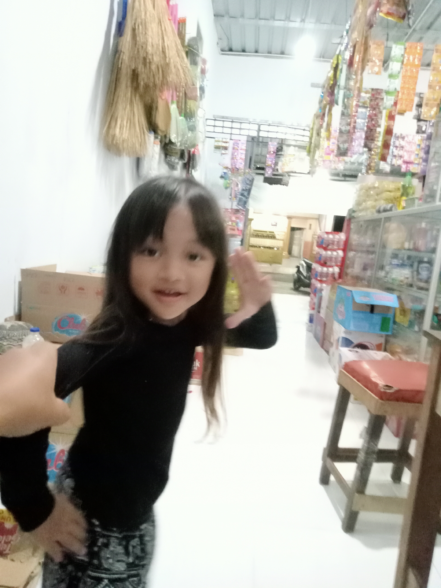 A young girl with long dark hair smiles at the camera. She is wearing a black long-sleeved shirt and patterned pants. She is standing in what appears to be a small shop or convenience store, possibly in Simo, Indonesia. Behind her, brooms hang on the wall, and shelves are stocked with various products. To her right, there are display shelves with stacked bottled water and other items. A wooden stool with a red cushion is visible in the foreground. The overall environment suggests a retail space with merchandise and cleaning supplies.