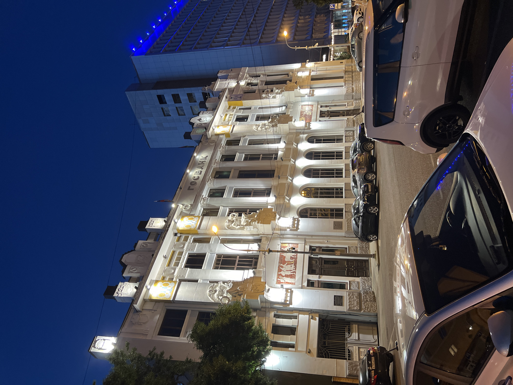 The image depicts a nighttime view of a building facade in Krasnodar, Russia. The building appears to be an older, ornate structure with white detailing, pilasters, and decorative sculptures. It is illuminated by spotlights, highlighting its architectural features. The text "ГОСБАНК" is visible on the building's facade, indicating it is likely a bank.
To the left of this building, a modern, taller building with blue lights is visible. Cars are parked along the street in front of the older building. The sky is a deep blue, characteristic of nighttime.
The scene is urban, with a mix of historical and contemporary architecture. The lighting suggests it is after dusk. No people are clearly visible. The overall mood is one of urban quietude at night, with the illuminated buildings standing out against the dark sky.