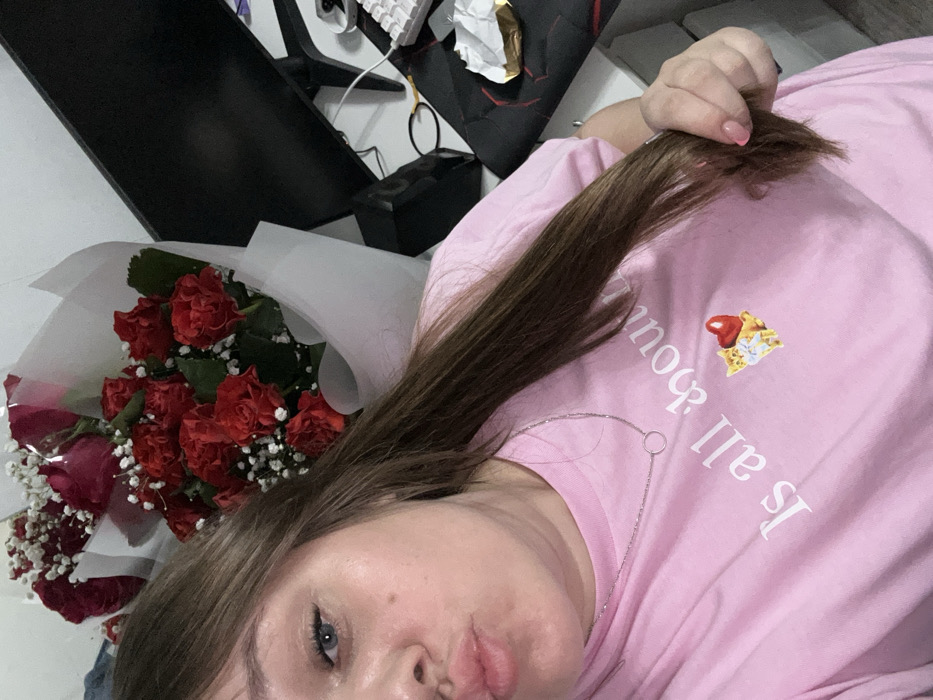 In an indoor setting within the city of Chaikovsky, Russia, a female individual is captured in a close-up, high-angle selfie. She has long, dark brown hair, blue eyes, and a pouting expression. Her right hand is visible, holding a portion of her hair, and her fingernails are painted with a light polish. She wears a light pink short-sleeved t-shirt bearing the partial text "Is all about" and a graphic depicting two small cats with a red heart. A thin silver chain necklace with a circular pendant adorns her neck. Behind and to her left, a large bouquet of red roses mixed with white gypsophila, wrapped in white paper, is prominent. To her right, a desk is visible, featuring a dark computer monitor, a white keyboard, a black object resembling a mouse pad, an open gold-colored wrapper, and a black hair tie.