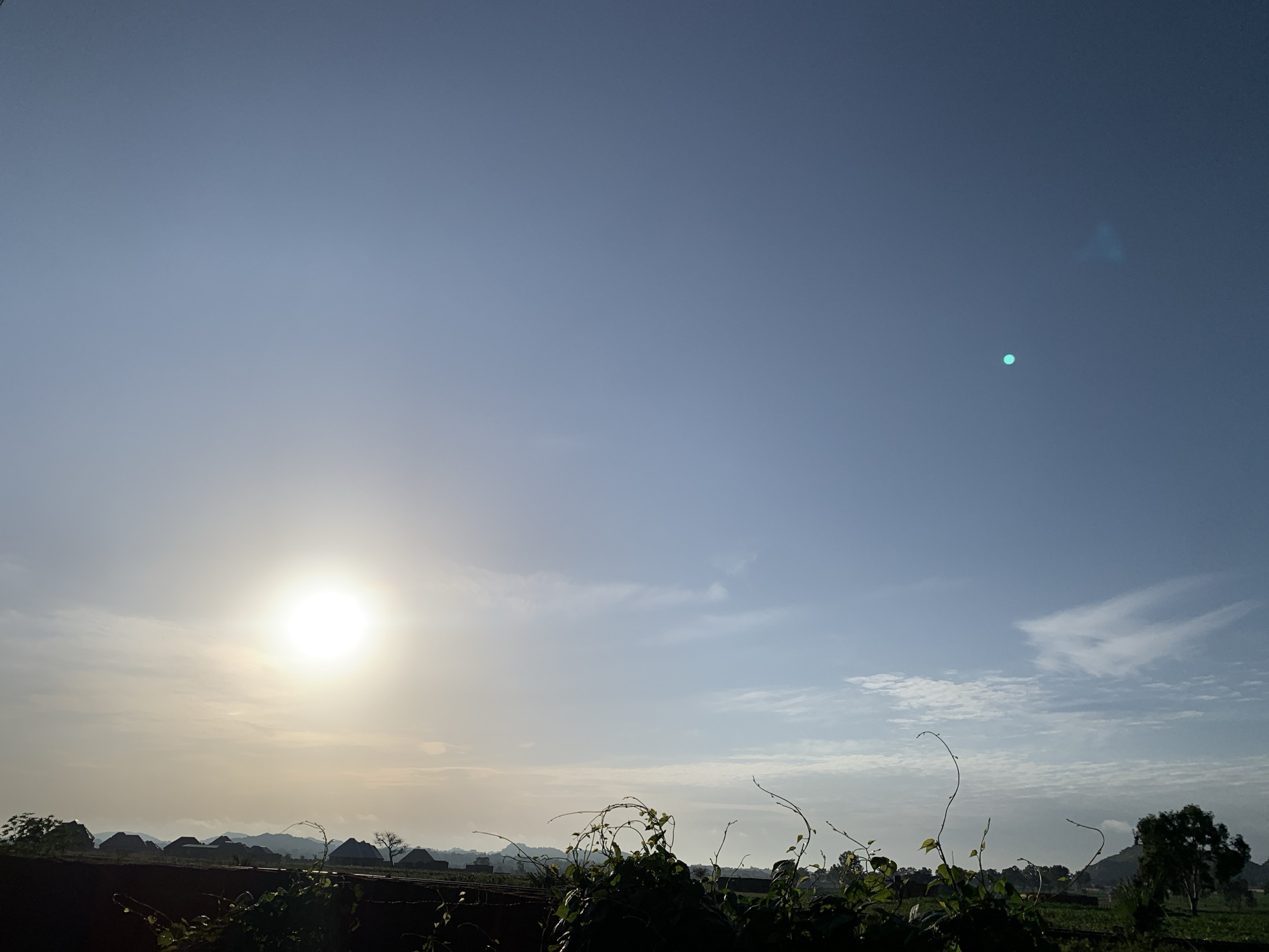 The image captures a wide-angle view of the sky over Kuru Karama, Nigeria, during what appears to be sunrise or sunset. The sun is a bright, luminous orb low in the sky, casting a warm glow that illuminates the surrounding atmosphere and creates a hazy effect over the distant landscape. The sky transitions from a bright yellow-white near the sun to a soft blue towards the top of the frame, with scattered wispy clouds. In the foreground, the silhouettes of lush green vegetation, including vines with delicate tendrils, are visible, contrasting with the hazy background. Beyond the vegetation, a line of thatched-roof structures, possibly traditional dwellings, are discernible against the backdrop of rolling hills or mountains in the distance. The overall impression is one of a serene rural setting bathed in the gentle light of the rising or setting sun.
