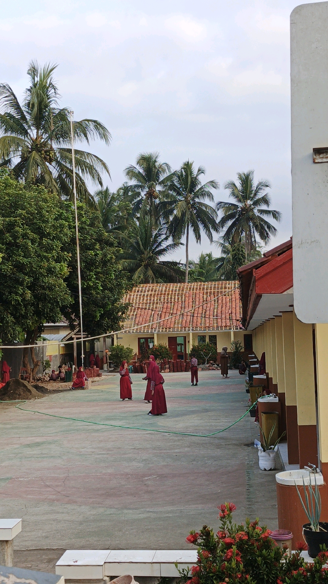The image depicts an outdoor scene at what appears to be a school or community center in Cibenda, Indonesia. Several individuals, predominantly wearing red attire, are present on a paved courtyard. Some are standing and conversing, while others are seated or walking. Coconut trees and lush green foliage frame the background, suggesting a tropical or subtropical environment. The architecture includes buildings with yellow walls and red tiled roofs, consistent with Indonesian building styles. A volleyball net is visible, indicating recreational activities might take place here. The overall impression is of a casual gathering or daily life in a warm climate.