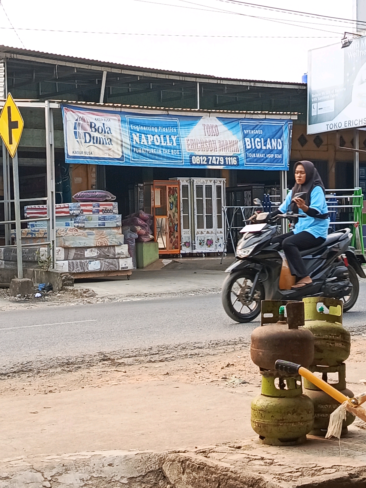 In Megang Sakti II, Indonesia, a woman wearing a black hijab and a bright blue shirt is riding a black motorcycle. She is looking down at her phone while riding. In the background, there is a storefront with a banner advertising mattresses and furniture. Several mattresses are stacked outside the store. In the foreground, two green propane tanks are visible. The scene appears to be a street in a commercial area.