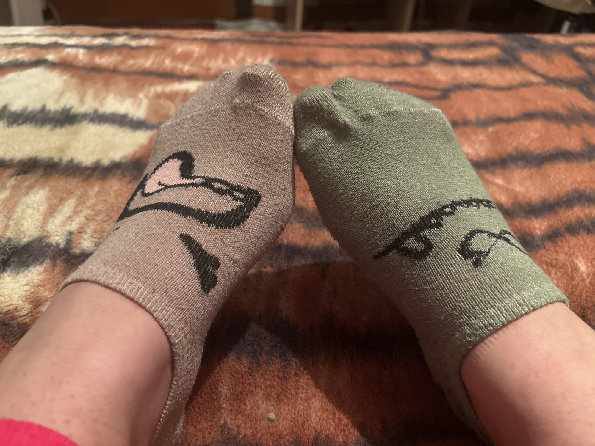 A pair of human feet, presumably belonging to one individual, are depicted resting on an animal-print blanket in an indoor setting within Dobrovnitsa, Bulgaria. The left foot wears a light brown ankle sock adorned with a black and pink stylized animal face design. The right foot wears an olive green ankle sock featuring a complementary black stylized design. Both socks are of a no-show style, exposing the ankles and lower portions of the feet. A small section of red fabric is visible near the left ankle. The blanket exhibits a prominent pattern of irregular stripes in shades of orange, cream, and dark brown/black, consistent with a tiger or similar animal print. The background is blurred, showing indistinct warm tones.