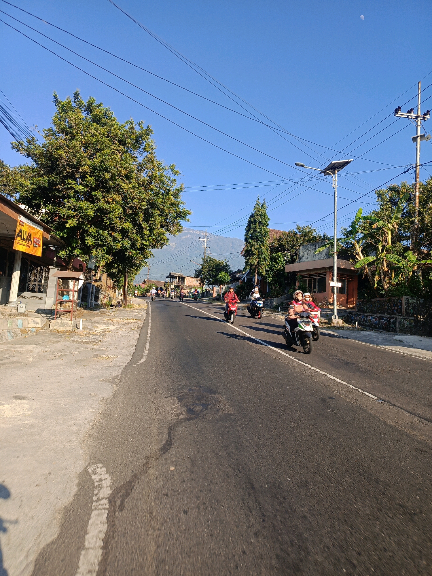 This is a daytime, eye-level, full shot photograph taken on a road in Kalang, Indonesia. The image depicts a rural road with a clear blue sky overhead. Several motorcycles with riders are traveling on the road. On the left side of the road, there are buildings and a large tree. On the right side, there are more buildings, trees, and a utility pole with electrical wires. In the distance, a mountain is visible under the sky. The overall scene suggests a sunny day in a residential area.