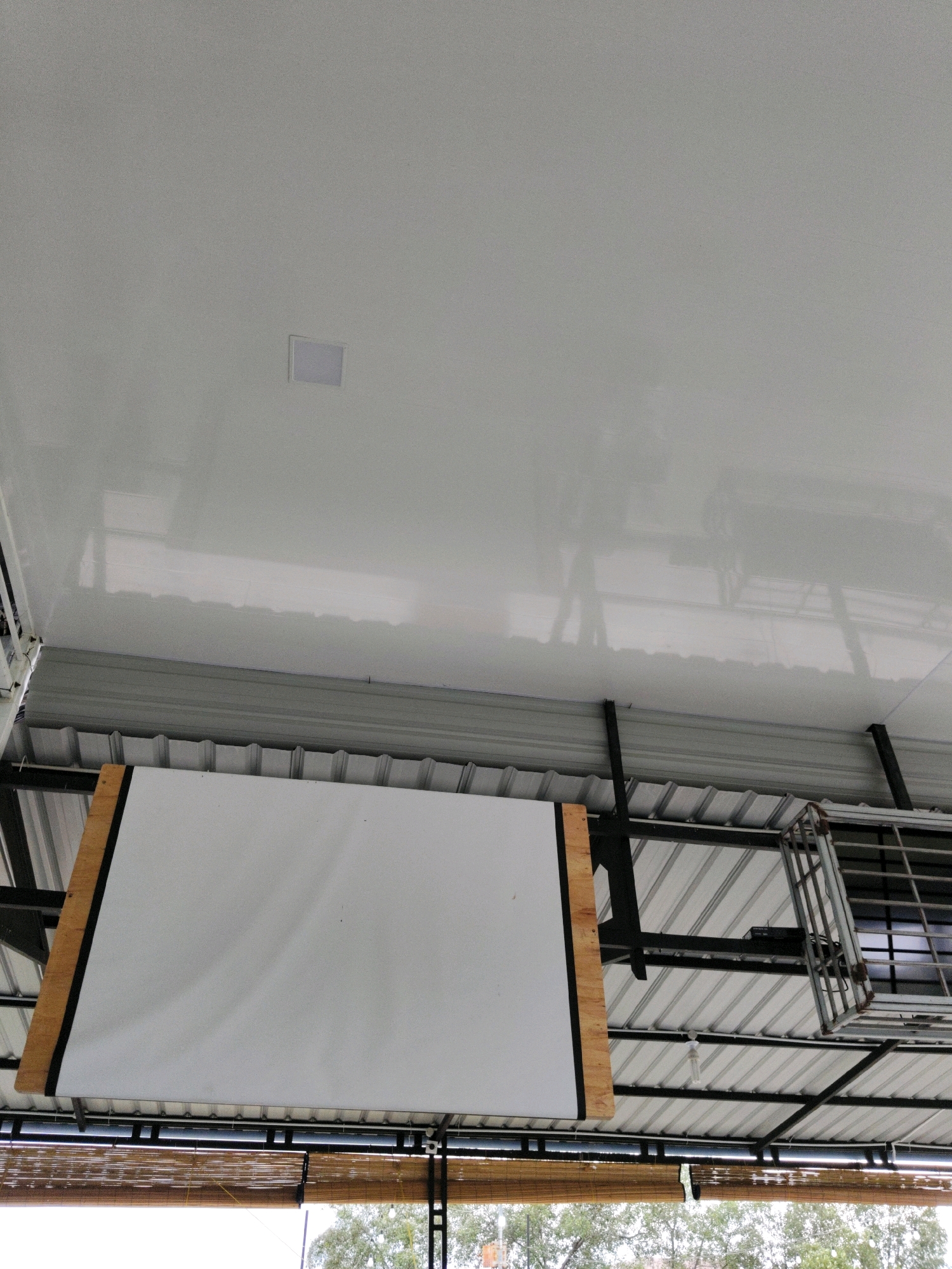 The image shows an interior view of a room, looking upwards at the ceiling and structures above. The ceiling is a smooth, white, reflective surface with a square light fixture in the upper central portion. Below the white ceiling, there is a layered structure. The first layer consists of gray, corrugated metal sheeting supported by black metal beams. Suspended from this structure is a large, white projection screen with wooden framing. Below the projection screen, a bamboo roller blind is visible. In the background, part of a metal rack with a grid pattern is present. The scene appears to be in an open-air or semi-open-air space, as trees and natural light are visible in the lower part of the image, suggesting it might be a covered patio or balcony. The presence of a projection screen implies the space could be used for presentations or entertainment. The overall setting is utilitarian and modern.