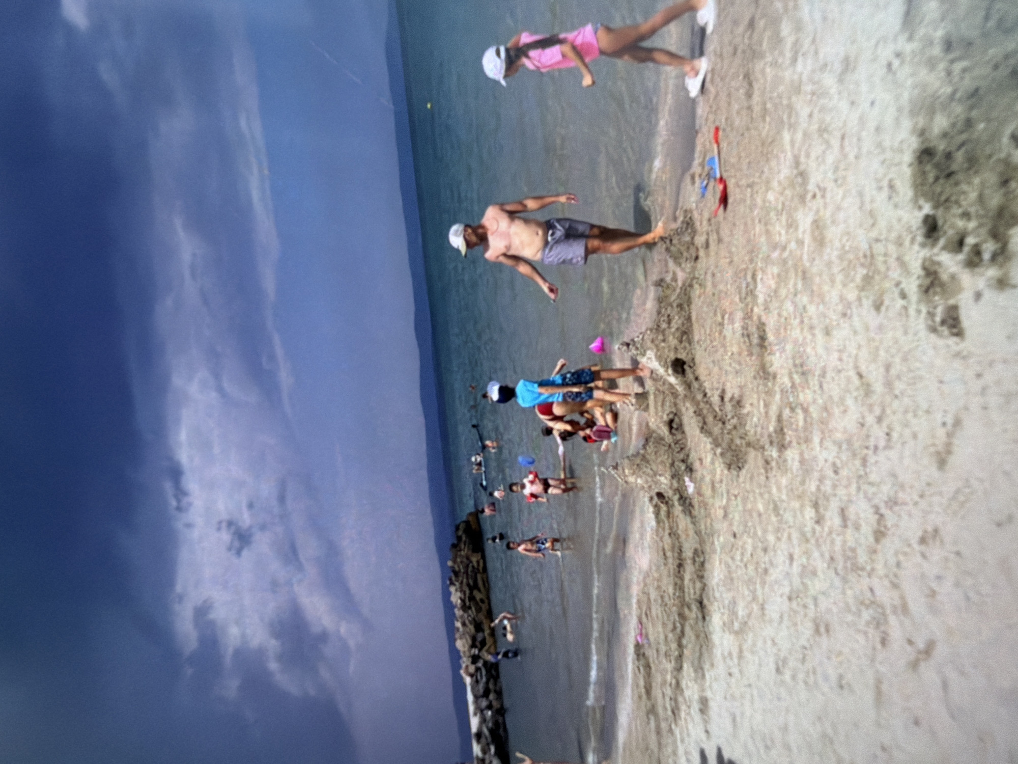 A wide shot captures a sunny day at a beach with a dark, stormy sky overhead. In the foreground, children and adults are engaged in various beach activities. A man in striped swim trunks and a white cap stands near the water's edge, facing a young girl in a pink swimsuit and white sandals. To their left, a boy in a blue swimsuit and a white cap is also in the water. The sand is damp and strewn with sandcastle remnants, and brightly colored toy shovels. Further back, other beachgoers can be seen swimming and playing in the shallow water. A stone breakwater extends into the sea on the left side of the image. The water is a clear turquoise, and distant hills form the horizon.