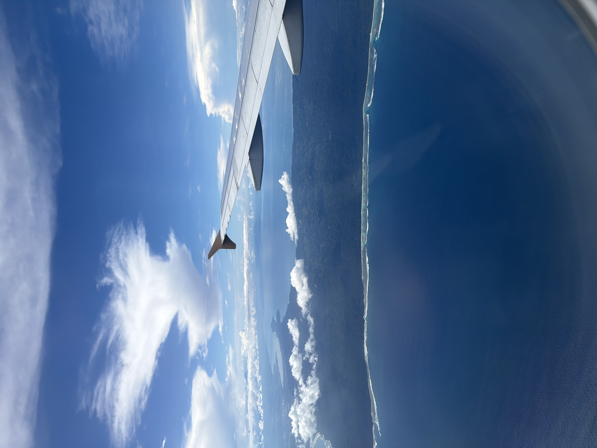An aerial view from an airplane window captures a vibrant tropical landscape under a clear blue sky. The image is oriented vertically, showing the airplane's wing extending diagonally from the top-left towards the center. The silver-grey wing, with its flap visible, occupies a significant portion of the upper-left frame.
Below the wing and extending across the middle of the frame is a lush, dark green landmass, indicative of dense vegetation, likely a coastline or island. This land appears to be partially covered by scattered white cumulus clouds. To the right of the land, a distinct, bright white line of breaking waves or shallow coral reefs marks the transition to the deep blue ocean, which fills the right side of the frame. A subtle reflection from the aircraft window is visible along the far right edge.
The sky is bright and expansive, dominated by a clear blue hue, with various formations of white clouds, including wispy cirrus clouds and more substantial cumulus clouds, scattered throughout. The time of day is bright daylight, suggesting good visibility and fair weather conditions. The combination of dense green land, clear turquoise shallows, and deep blue ocean, along with the distinct cloud patterns, strongly suggests a tropical coastal region. Given the provided context of Denpasar, Indonesia, the scene likely depicts a view over an Indonesian island or coastline. No people or visible text are present in the image.