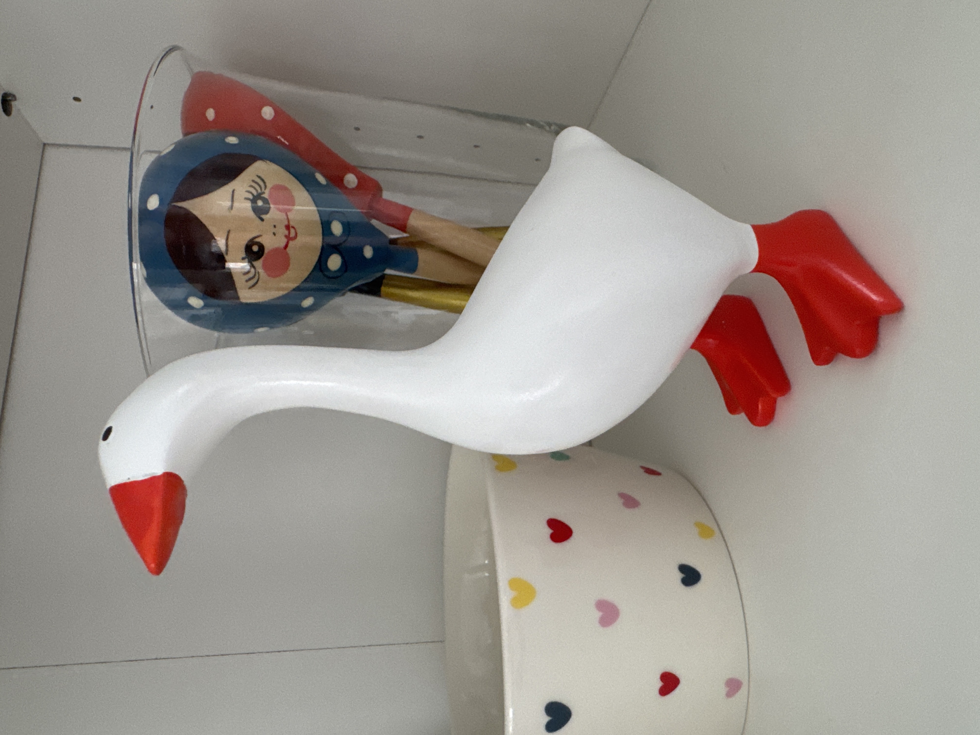 A white goose figurine with a red beak and red feet stands on a white shelf. To the left of the goose is a white bowl decorated with colorful hearts. To the right of the goose, a clear glass container holds wooden spoons. One spoon has a painted face resembling a matryoshka doll, and another is red with white polka dots. The shelf is likely in a home in Anudinovka, Russia, given the context.