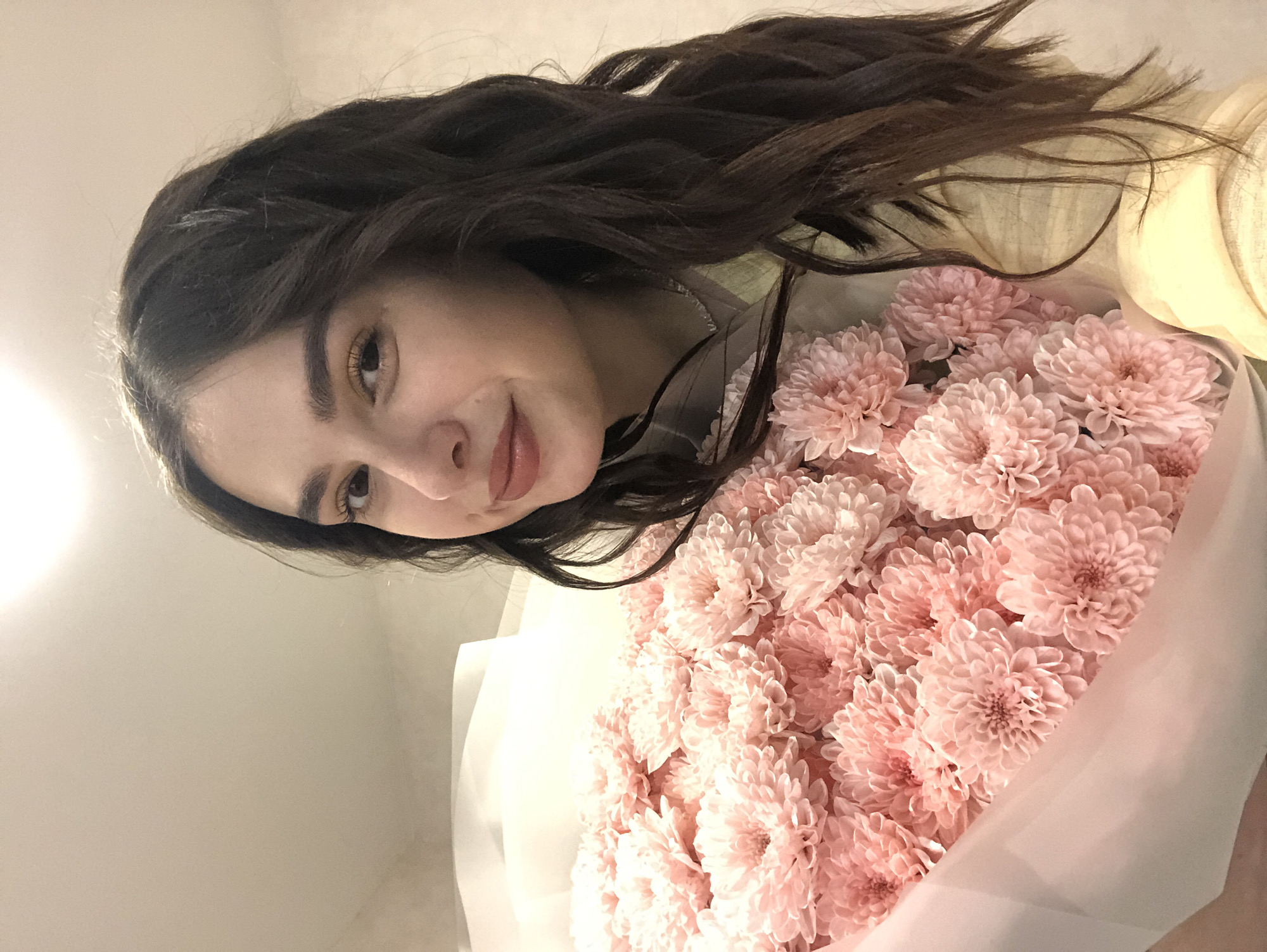 The image shows a young woman with dark, wavy hair, holding a large bouquet of pink chrysanthemums. She is wearing a light-colored top. The background is a neutral, light-colored wall, and a bright light source is visible at the top of the frame. The woman is looking towards the camera with a slight smile. The bouquet of flowers occupies a significant portion of the lower half of the image. There is no information in the image to indicate the location in Chaikovsky, Russia.