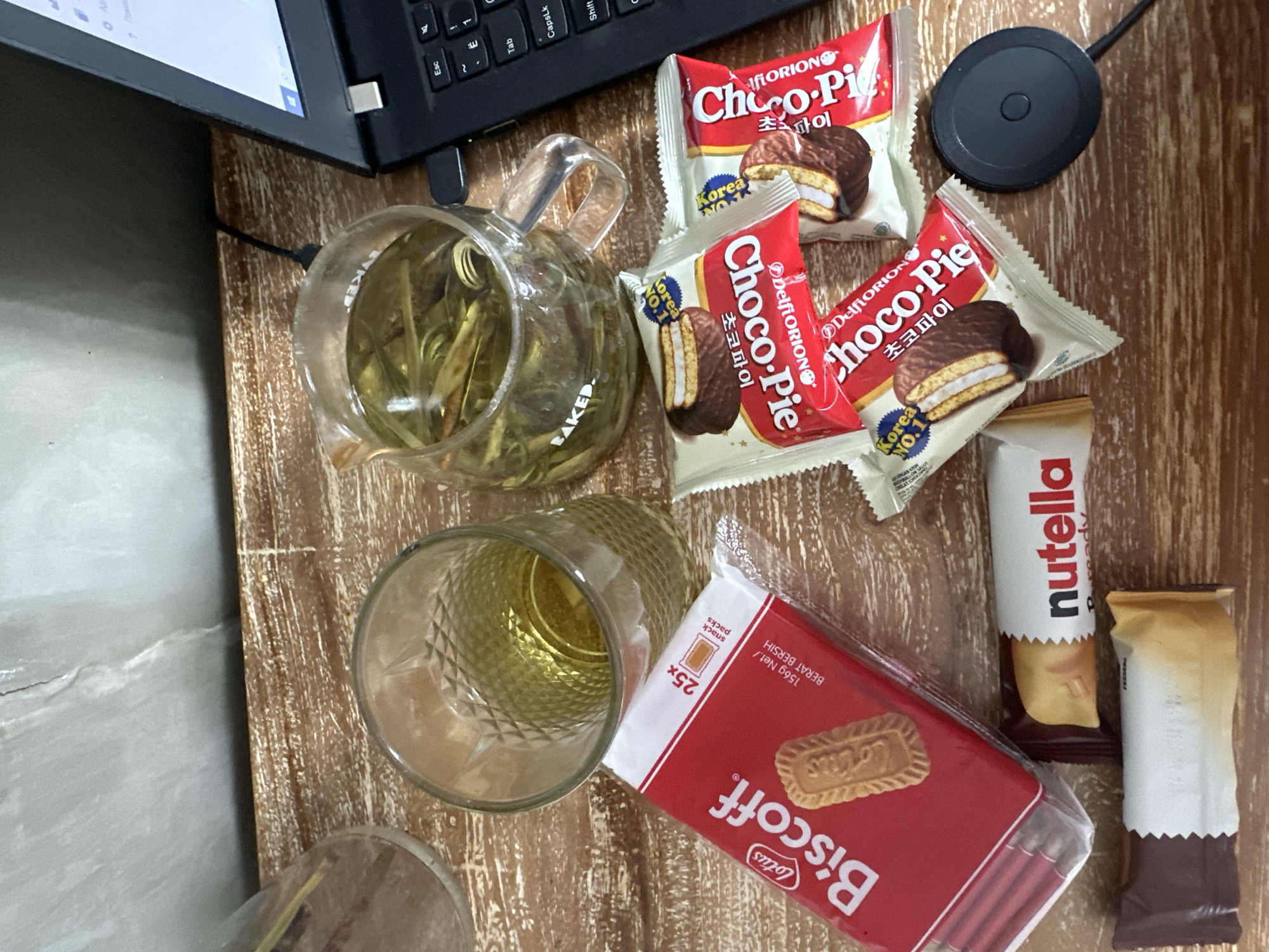 The image shows a top-down view of a wooden surface, likely a table, with a laptop partially visible at the top. On the table, there is a clear glass teapot with a handle, filled with a light-colored liquid and leafy contents. Next to the teapot is a tall glass containing a yellow liquid, possibly tea. Three individually wrapped Choco-Pie snacks, with red and white packaging, are also present. Adjacent to these are a Nutella snack and another individually wrapped snack with a white wrapper and brown accents. A rectangular package of Lotus Biscoff cookies, with a red wrapper, is located near the glass and the other snacks. A black, round wireless charger is positioned near the top right of the image. The setting appears to be indoors, possibly a workspace or a casual eating area. There is no evidence to confirm that the location is in North Kuta, Indonesia.