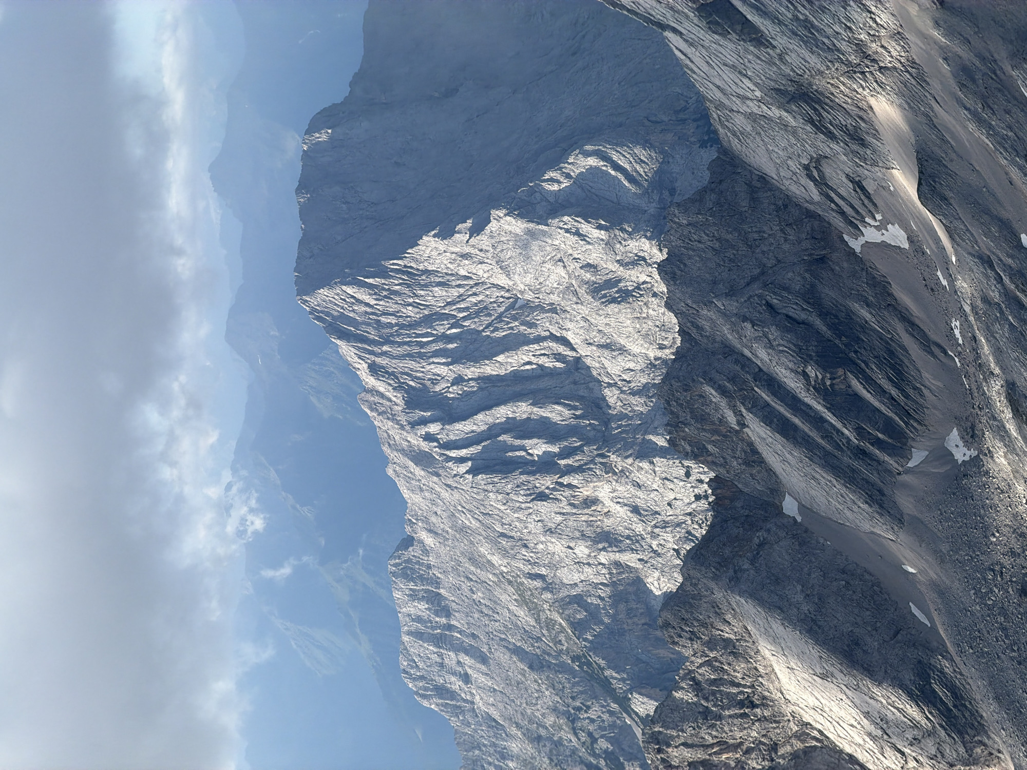 The image depicts a majestic mountain range, likely taken from a high vantage point overlooking Garmisch-Partenkirchen, Germany. The foreground is dominated by rugged, rocky mountain slopes characterized by steep inclines, sharp ridges, and shadowed crevices. Patches of snow are visible in shaded areas and lower elevations. The midground features a prominent, imposing mountain peak with a textured, weathered surface, catching the sunlight on its eastern face, creating stark contrasts of light and shadow. In the background, successive layers of mountains recede into the distance, gradually fading into a hazy, blue-toned atmosphere. The sky above is partly cloudy, with soft, diffused light filtering through the overcast conditions, suggesting either early morning or late afternoon. There are no people or man-made structures visible in the image, emphasizing the natural and remote character of the mountainous terrain.