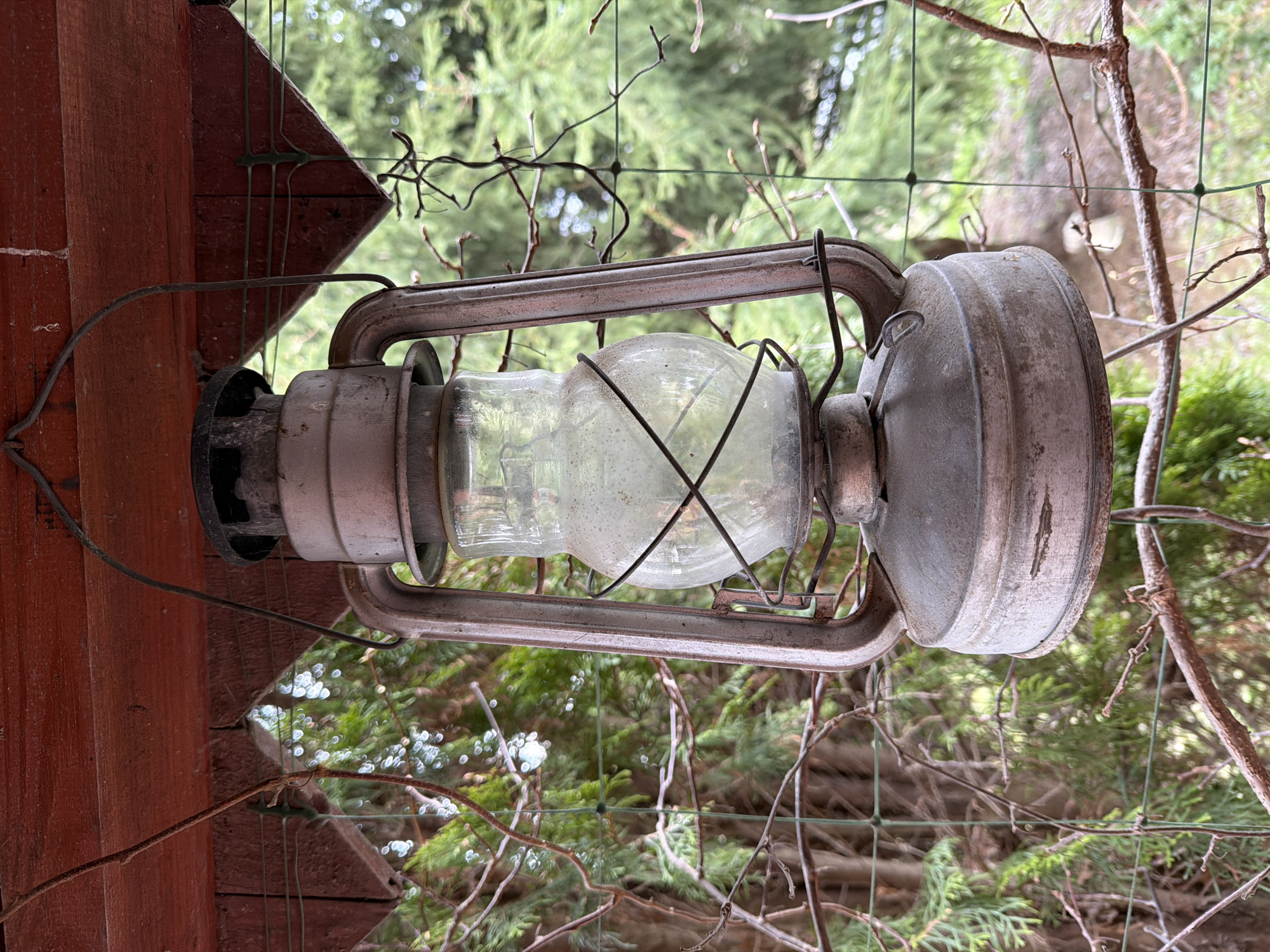 An old, weathered metal lantern hangs outdoors, its primary subject. The lantern features a dusty glass globe encased in a dark metal frame, with a rounded, patinated metal top and bottom. It shows signs of age and exposure to the elements, including scratches and dirt.
The lantern is suspended near a reddish-brown wooden structure, possibly a beam or post from a porch, pergola, or garden shed. A thin wire, likely aiding in the lantern's suspension or serving another purpose, runs along the wooden surface. Behind the lantern, a green wire mesh fence or trellis is visible, acting as a backdrop and separating the immediate foreground from the lush background.
The setting is outdoors, characterized by abundant green foliage, including coniferous trees and bare deciduous branches, suggesting a natural environment like a garden or backyard. The lighting indicates natural daylight, with soft illumination and some bokeh effects visible among the background leaves. No specific activity or event is taking place; the lantern is stationary, possibly serving as a decorative piece or historical artifact. The scene evokes a rustic atmosphere in an outdoor space within Пролетарский, Russia. There is no visible text in the image.