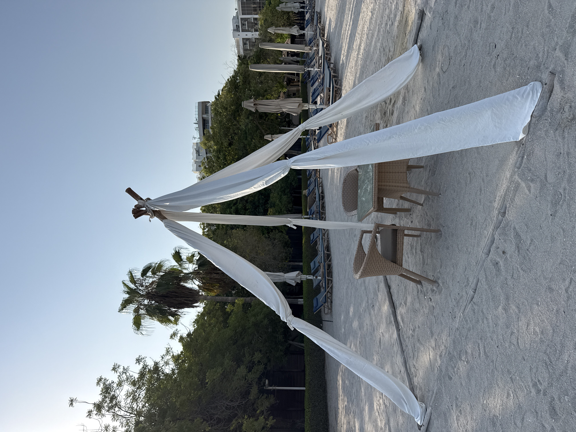 The image depicts a beach scene in Dubai, United Arab Emirates. In the foreground, a table with two wicker chairs is set up on the sand under a white canopy supported by wooden poles. The canopy drapes down, creating a sheltered area. Behind the table, rows of empty lounge chairs and closed umbrellas are neatly arranged on the sand, suggesting a resort or public beach area. Lush green trees and palm trees line the background, with several modern buildings visible in the distance. The sky is clear and blue, indicating a sunny day. There are no people present in the scene.