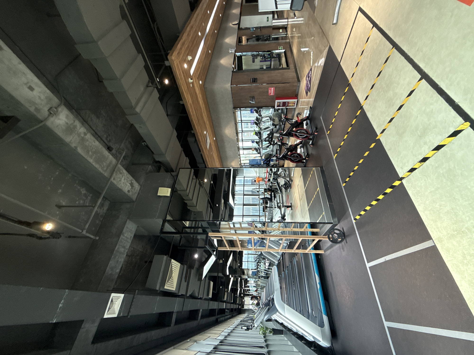 The image depicts the interior of a modern gym located in the Haizhu District, China. The gym features an array of exercise equipment, including numerous treadmills lined up along a large window overlooking a cityscape. Several stationary bikes are positioned in the foreground. The gym's architecture includes an exposed ceiling with visible ductwork and lighting fixtures. Mirrors line one wall, reflecting the equipment and the overall space. The floor is a mix of dark grey rubber matting with yellow and black hazard tape marking designated areas, and lighter colored flooring. A wooden ladder structure is visible in the mid-ground. A few individuals are present in the gym, engaged in exercise or moving around the space. The environment appears well-lit and organized.