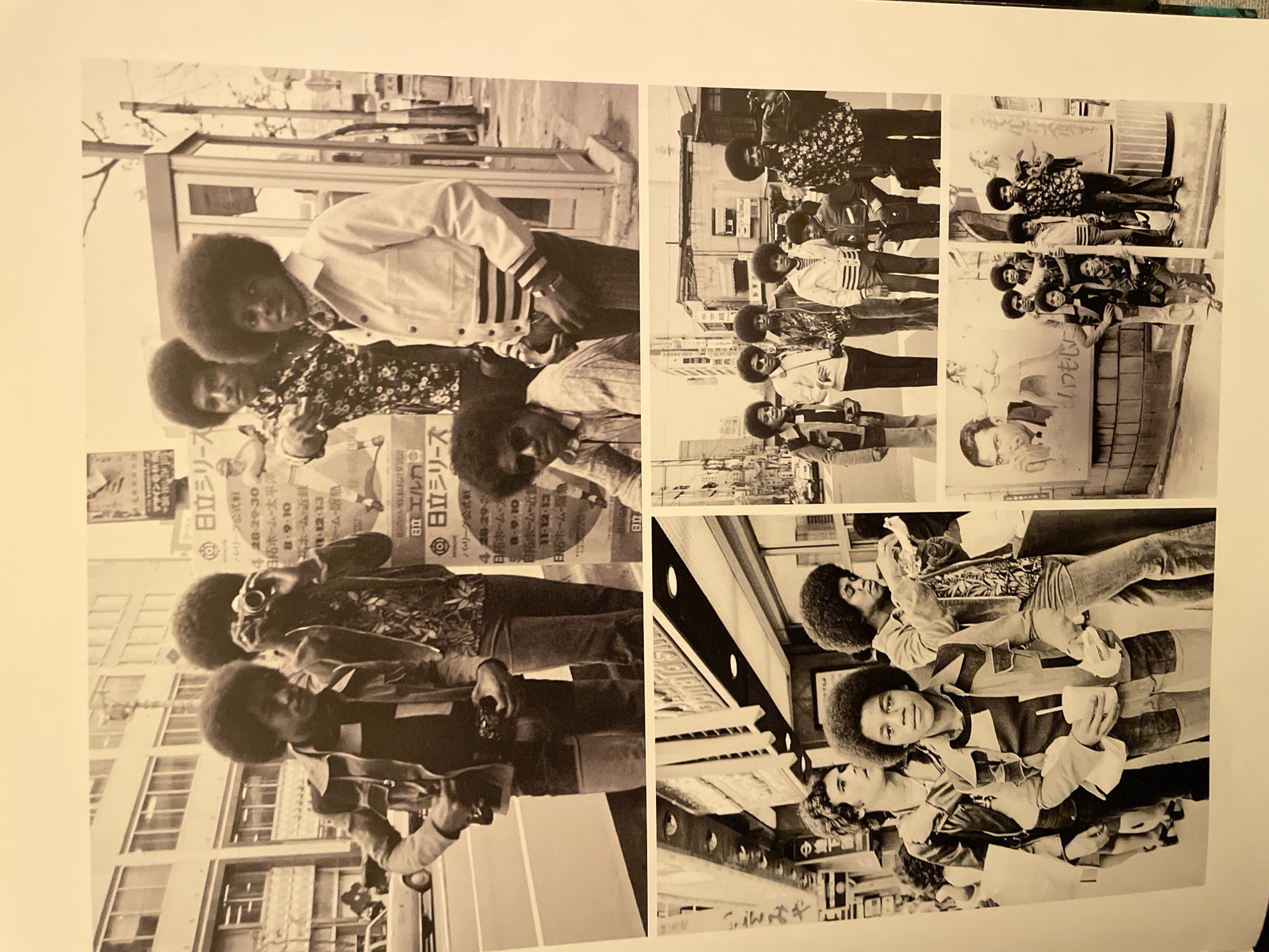 This image presents a collage of six black and white photographs, primarily featuring a group of five young men, identified as the Jackson 5, posing in various urban settings. The visible architecture, street signs, and prominently displayed Japanese characters on posters and storefronts indicate the location is a city in Japan, not Barcelona, Spain.
The top, largest photograph captures five members of the Jackson 5 on a city street during the day. All five men wear distinct 1970s attire, including button-up shirts, jackets, and bell-bottom-style trousers, with prominent afro hairstyles. From left to right: one man in a patterned shirt and jacket holds a camera, looking down. Next to him, a man with a camera to his eye appears to be taking a photo. Behind them, another man in a dark, floral-patterned shirt points towards the right. To his right, a man in a light-colored jacket and patterned scarf poses with his hands in his pockets. Seated in the foreground, below the pointing man, is another individual wearing sunglasses and a patterned shirt. In the background, there are multi-story buildings, a parked white car, and a public telephone booth. Numerous signs with Japanese text are visible, including those for "日立" (Hitachi) and numeric sequences suggesting dates (e.g., "4.28-29.30").
The bottom section of the image contains five smaller photographs.
The bottom-left photo features four men from the group standing on a sidewalk in front of a building with an awning. A light-skinned man with curly hair stands at the far left. The central figure, Michael Jackson, stands front and center, holding a plastic cup with a straw and a wrapped food item in his hands. He wears a multi-paneled jacket over a striped shirt. Another group member is seen to his right, wearing a patterned shirt.
The remaining four smaller photos, located on the bottom right, depict the group in similar street environments. One photo shows several members posing in front of a wall with Japanese posters, one of which depicts a man's face. Another shows a man carrying a young child on his shoulders, with other people and storefronts in the background. The clothing styles and general urban environment remain consistent across all depicted scenes.