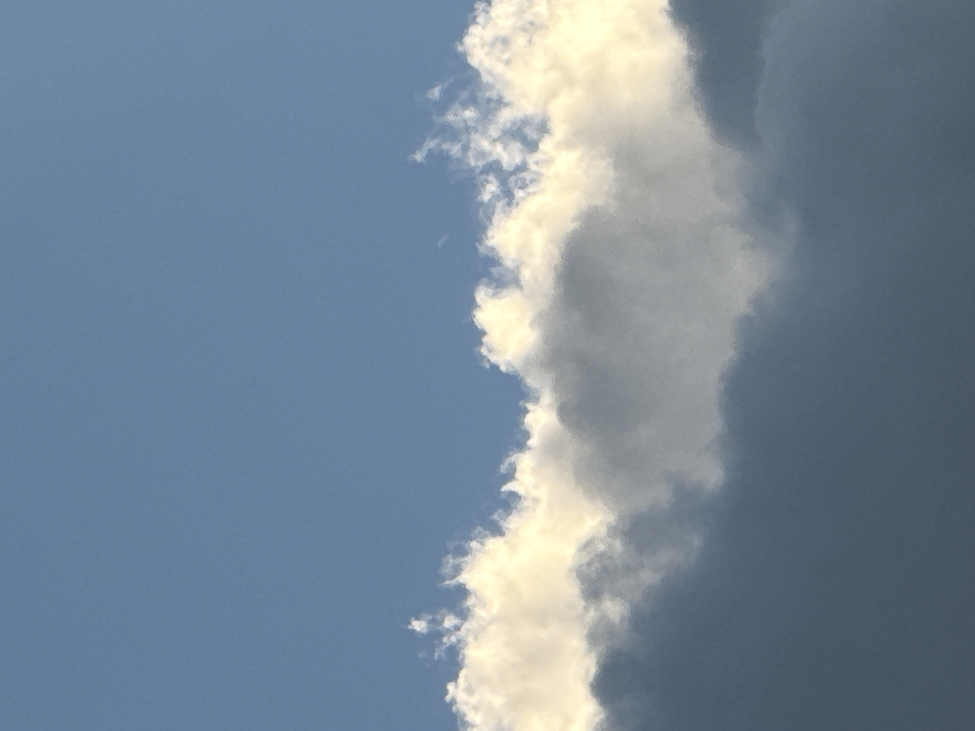 The image depicts a portion of the sky above Voeikovo, Russia. The upper half of the image is dominated by a clear, blue sky. The lower half features a layered arrangement of clouds. The topmost layer of clouds is wispy and white, with some appearing illuminated with a yellowish hue, suggesting sunlight. Below these, a thicker, gray cloud formation extends across the frame, with its upper edge showing some lighter, textured areas where it meets the brighter clouds. The lowest visible portion of the image is a darker, more solid gray cloud layer. The scene is devoid of any human presence, structures, or discernible objects, focusing solely on the atmospheric conditions.