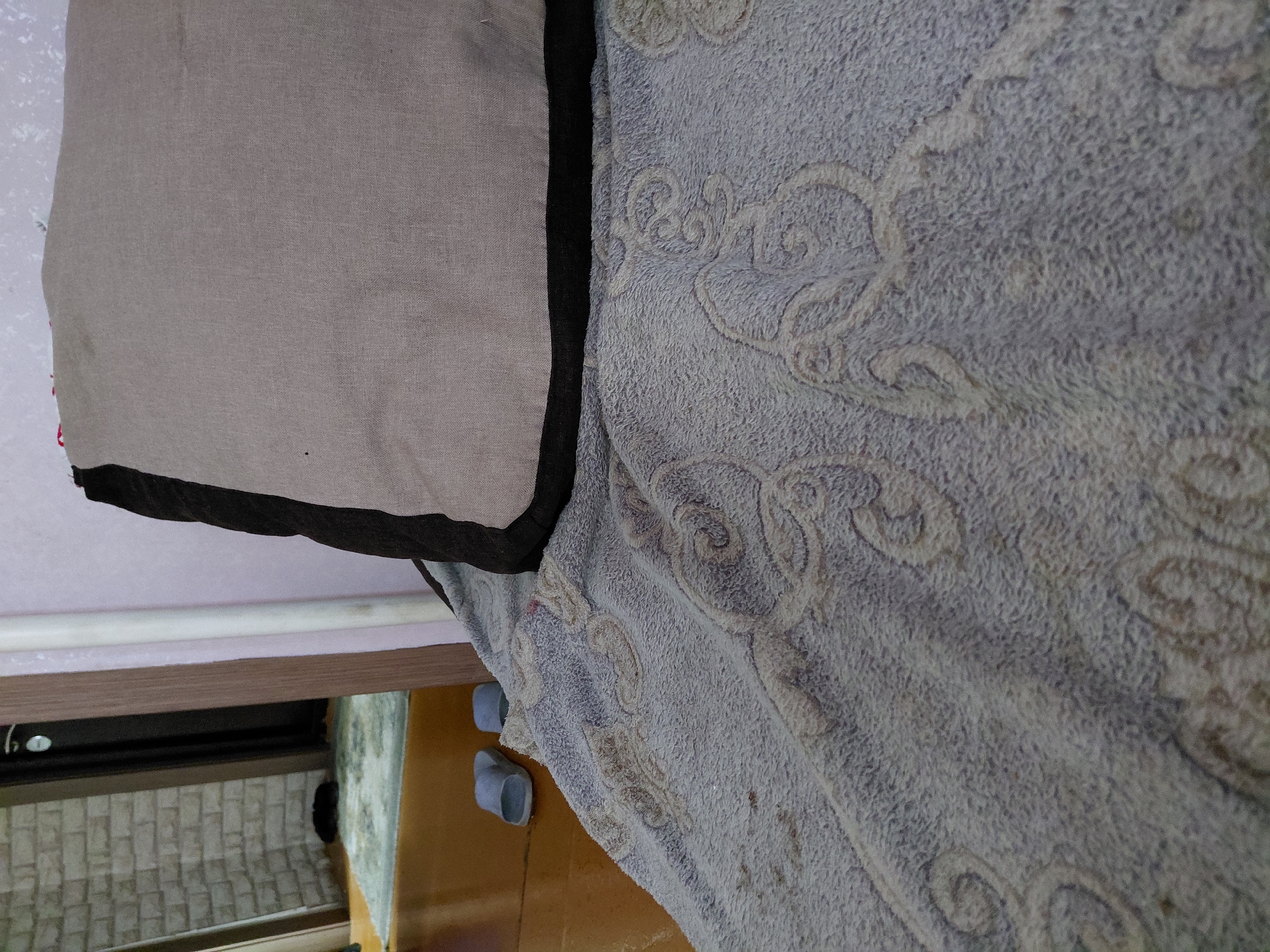 In Malinovka, Russia, an indoor scene focuses on a close-up view of a bed or sofa covered with a textured, light blue blanket featuring an ornate, swirling pattern. A light beige pillow with a dark trim is placed on top of the blanket, partially obscuring the pattern. In the background, a doorway leads to another room, where a brick wall is visible. A pair of light blue slippers rests on a wooden floor in the background. The overall impression is of a domestic setting, likely a bedroom or living area.