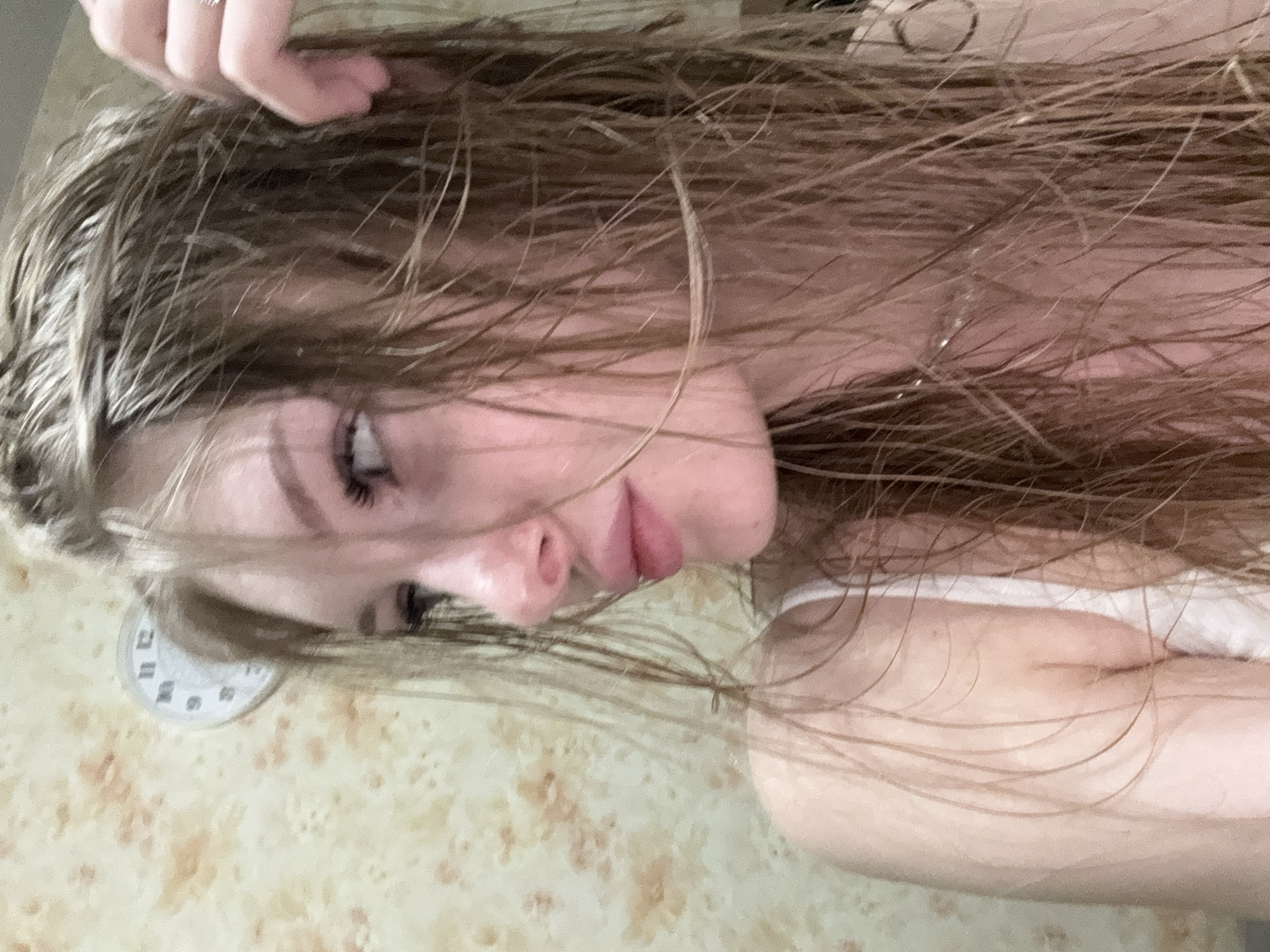 The image displays a young individual, likely female, captured in a left profile view, looking towards the left side of the frame. Her long, light brown hair appears wet and somewhat disheveled, falling across her left shoulder and chest, and partially obscuring parts of her face. Her right hand, adorned with a simple metallic ring on the ring finger, is visible in the upper right portion of the frame, gently touching or running through her hair near her right temple. She wears a white top with a visible strap on her left shoulder. Her visible facial features include prominent, possibly enhanced, eyelashes, a softly defined eyebrow, a small nose, and softly closed or slightly pursed lips. The background consists of a light-colored wall, possibly covered in wallpaper, featuring a subtle, irregular, mottled pattern in shades of cream and light orange-brown. Partially visible behind her head in the upper left quadrant is a circular white wall clock, showing portions of the numbers 9, 11, 12, and 1. There are no discernible visual elements within the image to identify its specific location as Perm, Russia.