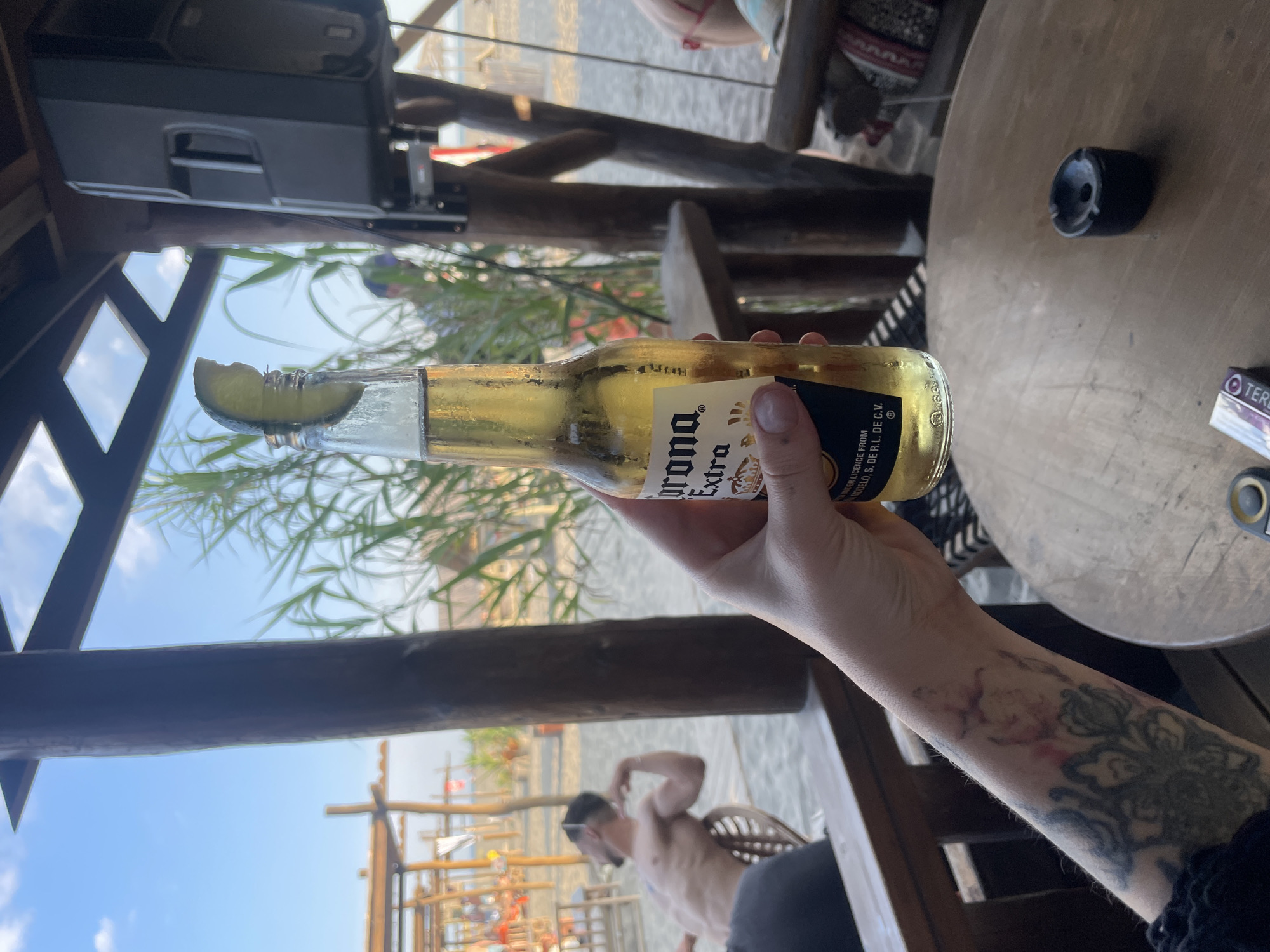 A hand holds a bottle of Corona Extra with a lime wedge in its neck, positioned in the foreground. The bottle is partially submerged in a liquid. Behind it, a man with dark hair and sunglasses is visible from the chest up, shirtless, sitting on a chair and looking to his left. He has tattoos on his back. The background suggests an outdoor setting, possibly a beach bar, with wooden structures, vegetation, and sand. A round wooden table with an ashtray and cigarette pack sits in the lower right corner. The overall lighting indicates daytime. The scene is located in Donji Štoj, Montenegro.