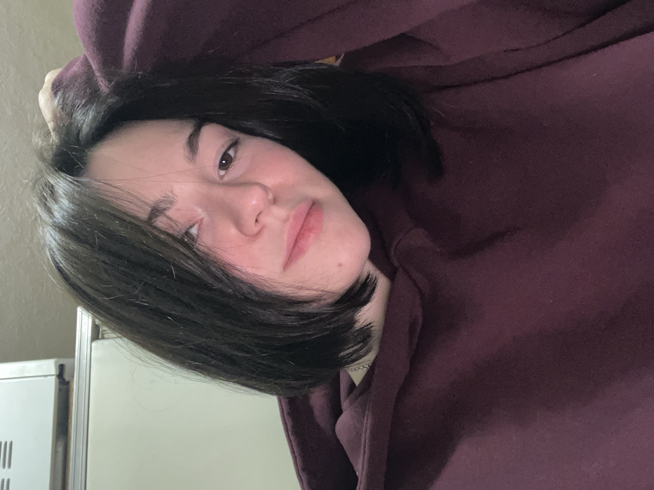 A young woman with dark brown hair styled in a short bob, wearing a maroon hooded sweatshirt, gazes towards the camera with a slight smirk. Her complexion appears fair with rosy cheeks. Part of a white appliance, possibly a refrigerator or oven, is visible in the background on the left side of the frame. The overall image is angled, creating a casual and relaxed vibe.