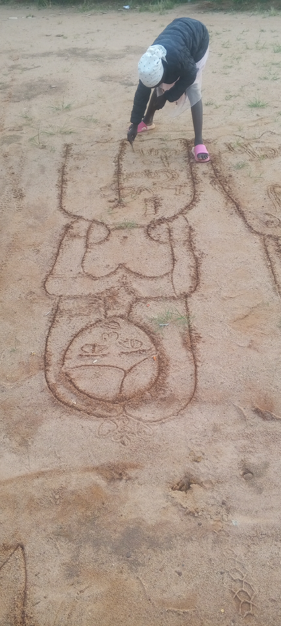 A person, wearing a dark jacket, a light-colored headscarf, and pink sandals, is drawing a figure in the sand. The drawing appears to be a large, stylized human form with distinct sections representing the head, torso, and lower body, possibly depicting a pregnant individual. The person is using a stick or a finger to outline the figure in the sandy ground. The environment is outdoors, with sparse grass and uneven terrain, suggesting a rural or undeveloped area. The sand is a light brown color. The overall scene is simple and focused on the act of creation in the natural environment.
