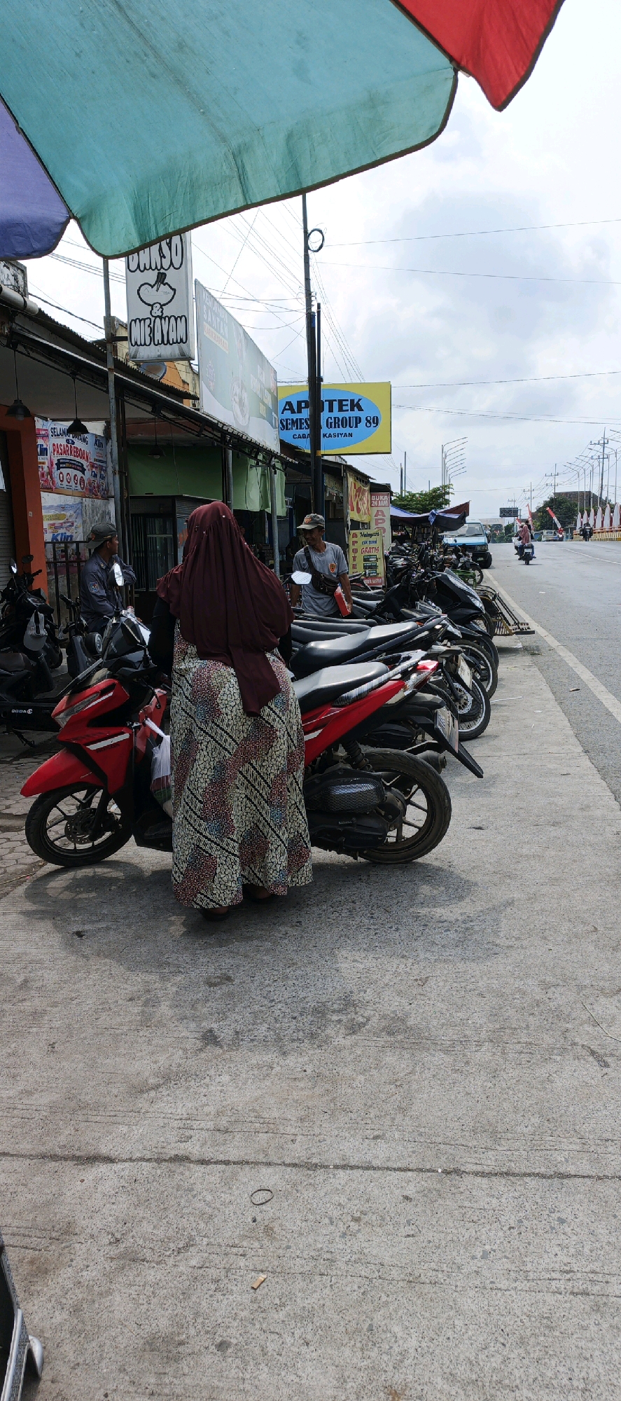 The image depicts a street scene in Kasiyan, Indonesia, with a focus on a row of parked motorcycles along a sidewalk. A woman wearing a dark head covering and a patterned long skirt stands next to a red motorcycle, partially obscuring it. To her left, two men are visible, one sitting on a motorcycle and wearing a grey uniform with a cap, and another standing and wearing a grey t-shirt and a cap, holding a red object. Behind them, a shop facade with various signs is visible, including one for "DURSO MIE AYAM" and another for "APOTEK SEMES GROUP 89". The environment includes a paved road with more motorcycles and traffic in the background, overhead power lines, and a large, colorful awning. The sky is cloudy.