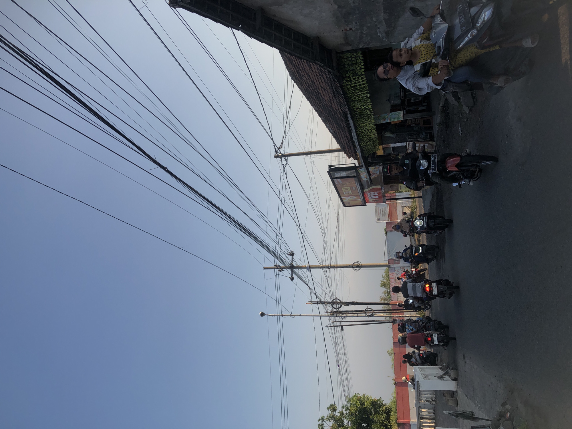 This is an eye-level, outdoor, full shot taken on a street in Gempolsari, Indonesia. The sky is clear and pale blue, with many overhead power lines stretching across the frame. A few bare trees are visible on the left side of the image, and a building with a tiled roof and a grey, weathered facade is on the right. The street is paved with asphalt, and there are several motorcycles with riders on it, moving in both directions. The people are casually dressed, and the overall atmosphere appears to be that of a typical day in a residential or commercial area.