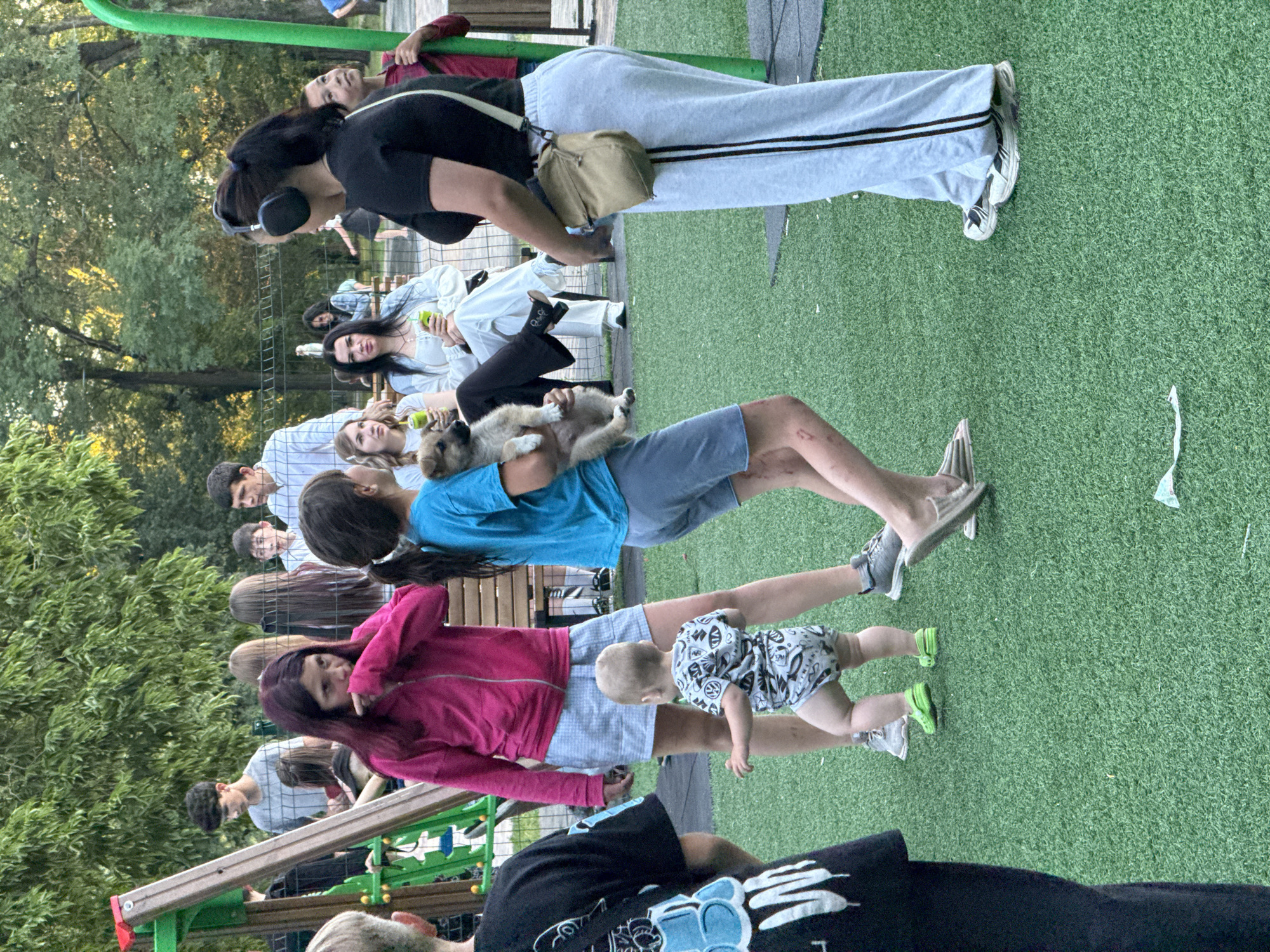 A group of people are gathered on a bright green turf area, likely a park or playground, with trees in the background. In the foreground, a young child wearing a patterned onesie is standing and appears to be reaching out, with green shoes on. To the left of the child, a young girl in a blue t-shirt is holding a small brown puppy. Behind them, a woman in a bright pink zip-up hoodie and a checkered skirt stands with her hands near her mouth. Several other individuals, including adults and children, are visible in the middle ground and background, some sitting on benches and others standing. The lighting suggests it is daytime, possibly late afternoon, with sunlight filtering through the trees. The overall scene depicts a casual outdoor gathering.