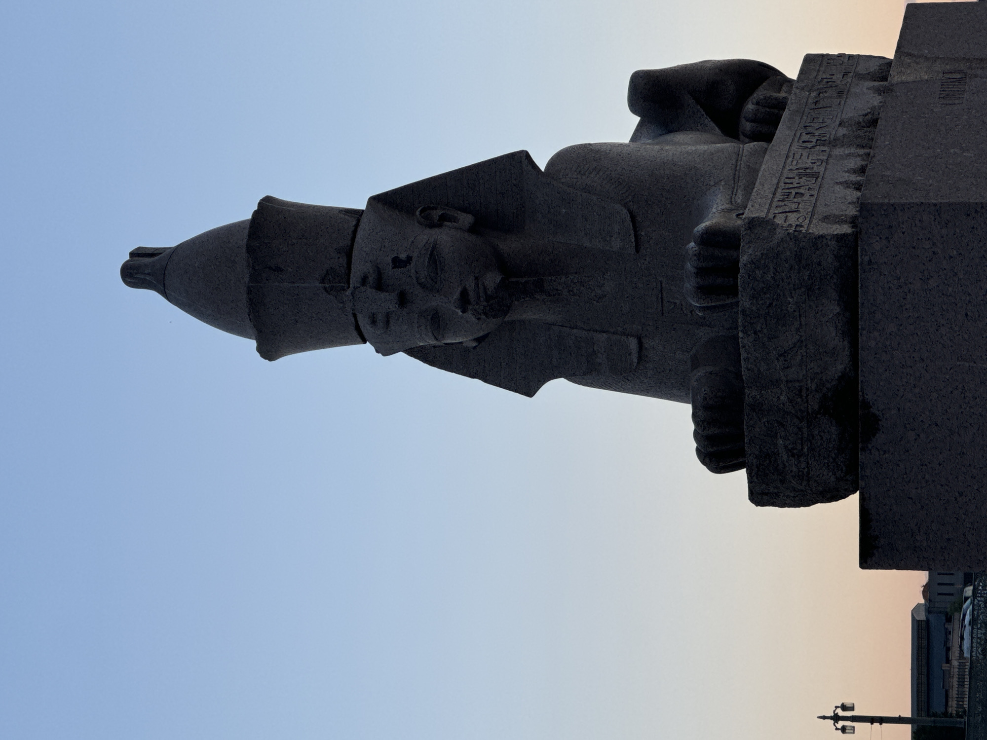 The image shows a stone sphinx statue in Saint Petersburg, Russia, against a clear sky at dusk. The sphinx is a dark gray, ancient Egyptian-style statue with a human head and a lion's body. It is seated on a granite pedestal inscribed with hieroglyphs. The statue is illuminated by the soft light of the setting sun, casting long shadows. In the background, a streetlamp and the outlines of buildings are visible. The overall mood is serene and imposing, conveying a sense of history and timelessness.
