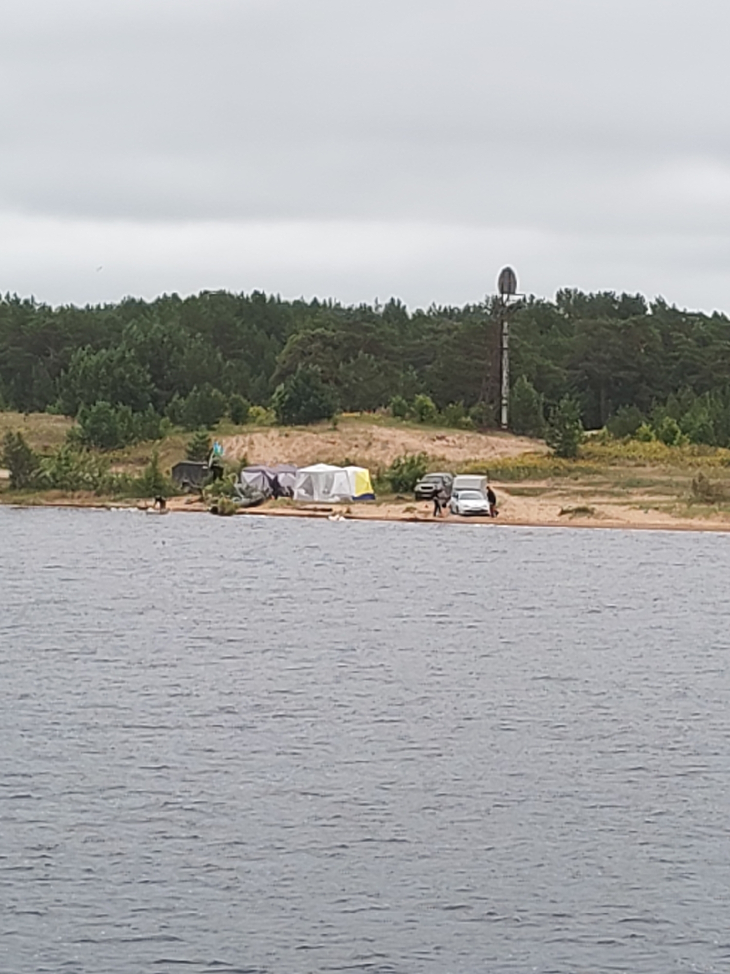 The image depicts a campsite on a sandy shore adjacent to a body of water. A line of pine trees forms the background, with a cloudy sky above. Several tents are set up on the beach, including white, grey, and a yellow and blue striped tent. Three vehicles are parked near the campsite: a dark SUV, a white camper van, and a white sedan. Several people are visible, some standing near the vehicles and tents, and at least one person is in the water. A tall, slender tower with a spherical top stands on the right side of the image, behind the trees. The overall scene suggests an outdoor recreational activity, possibly camping or fishing, in a natural environment.