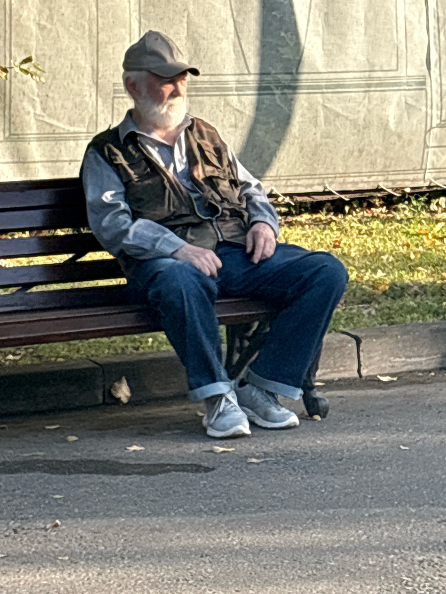 An elderly Caucasian man with a white beard and a gray baseball cap sits alone on a park bench. He wears a light blue long-sleeved shirt, a dark olive vest with multiple pockets, and dark blue jeans that are cuffed at the ankles. His feet are clad in light gray sneakers. The man appears to be looking off to his right. He is seated on a dark brown wooden bench. Behind the bench, there is a large, light-colored tarp or backdrop with horizontal lines. To the right of the bench and behind it, a patch of green grass is visible with scattered autumn leaves. The foreground consists of a paved pathway with some fallen leaves and a dark, irregular shadow cast from an unseen object. The overall impression is of a tranquil, sunlit park setting.