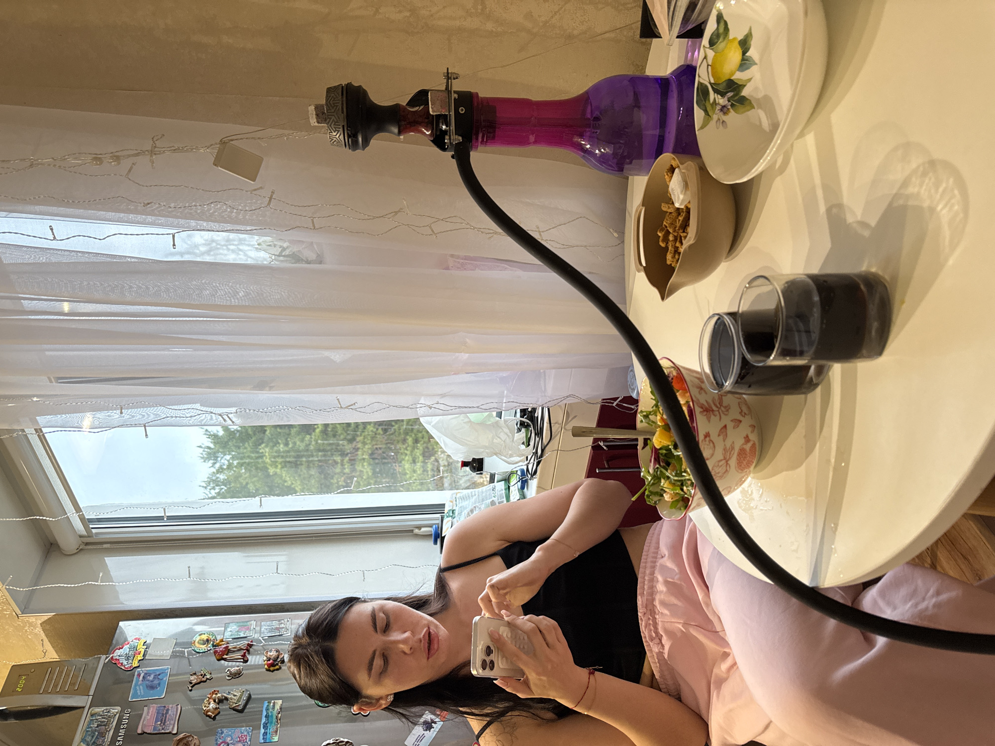 A woman is taking a photo with her phone in a room. The scene includes a table with dishes, a hookah, and a window offering a view of trees. The location appears to be in Chaikovsky, Russia, based on a magnet visible on the refrigerator.