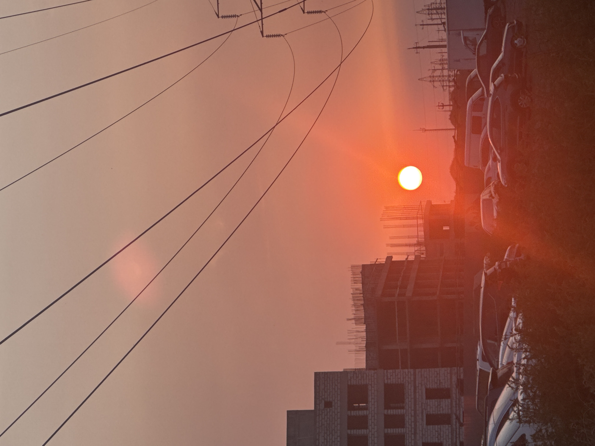 The image depicts an outdoor scene at sunset, possibly in an urban or industrial area. The sky is a vibrant orange-red, indicating the sun is low on the horizon. Several power lines are prominently strung across the frame, some appearing taut and others with a slight sag. In the foreground, parked cars are visible, mostly obscured by foliage. Behind the cars, there are unfinished or under-construction buildings with exposed rebar. In the background, a series of utility poles and structures, possibly part of a power grid or industrial facility, are silhouetted against the sky. The overall atmosphere is one of a city at dusk, with the natural beauty of the sunset contrasting with the man-made structures.