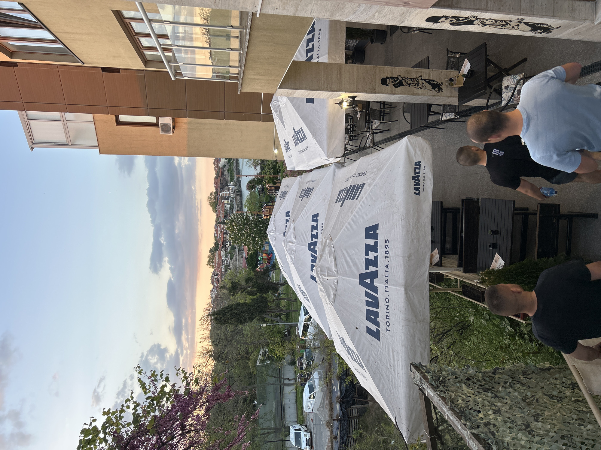 This eye-level outdoor shot presents a view of a restaurant in Sozopol, Bulgaria. The shot is oriented in a vertical fashion. Several white canopies emblazoned with the "LAVAZZA TORINO, ITALIA, 1895" logo are prominently featured, suggesting that the restaurant may serve Lavazza coffee. Tables and chairs are placed underneath the canopies.
In the foreground, there are two men who are only partially visible. One is wearing a blue shirt and is slightly out of focus, while the other one is wearing a black shirt.
The background shows a cityscape with buildings of various heights and designs. The sky is partially visible, displaying a gradient of colors from light blue to pale orange, hinting at either dawn or dusk. The surrounding environment includes trees, shrubs, and patches of greenery.
The overall atmosphere of the scene appears to be calm. The restaurant seems to be getting ready for service, or perhaps in the midst of it. The urban setting provides a contrast to the natural elements of the sky and greenery.
