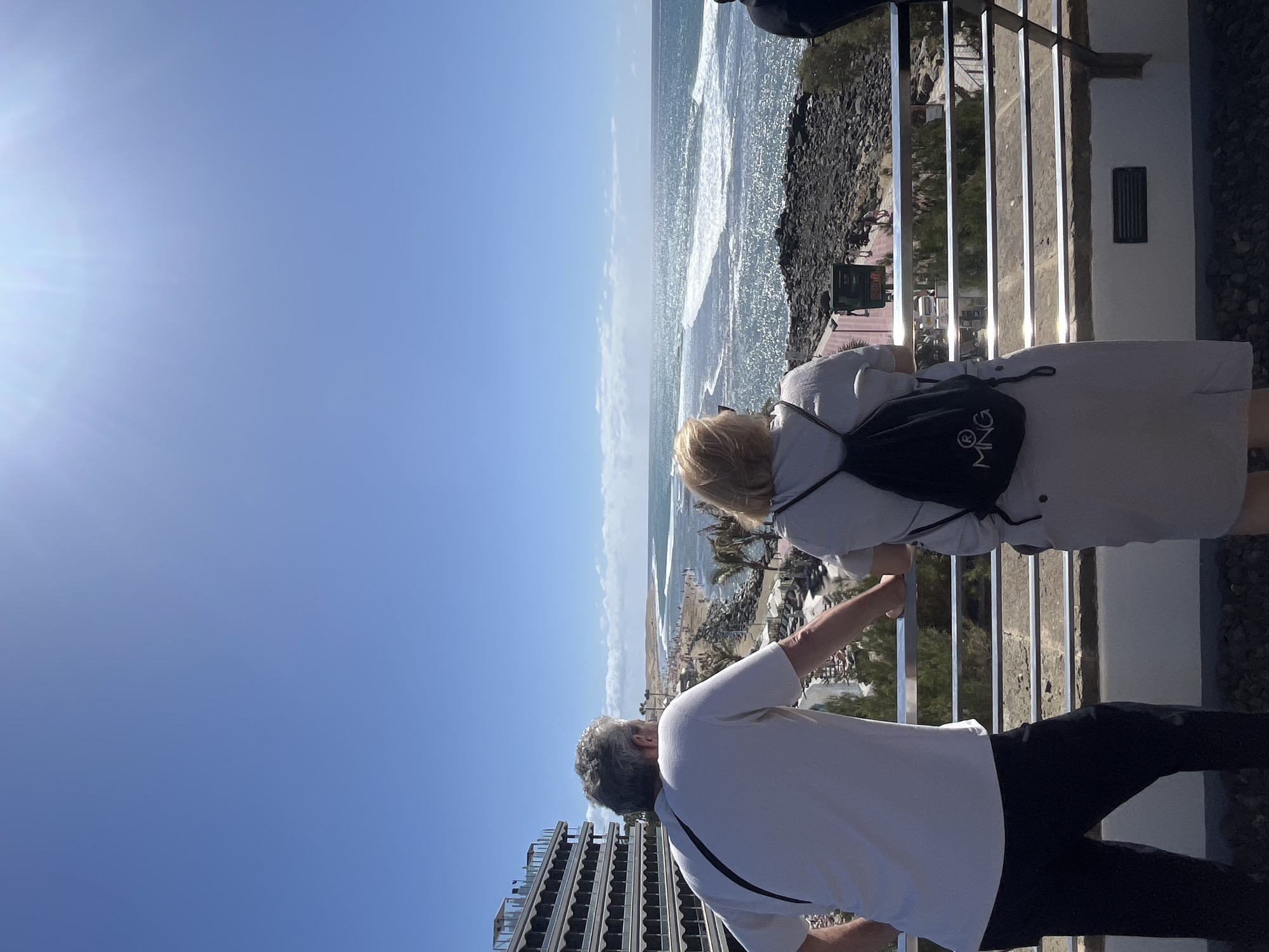 Two individuals stand behind a silver metal railing, looking out at a coastline. The person on the left, presumably male, wears a white t-shirt and black pants, and points with his right hand. The person on the right, presumably female, has blonde hair and wears a light-colored dress with a black backpack. The background shows a sunlit ocean with crashing waves, a beach with palm trees, and a rocky shoreline. To the left, a modern building with a stacked balcony design is visible. Given the context of Maspalomas, Spain, the location may be a hotel balcony or a public overlook point.