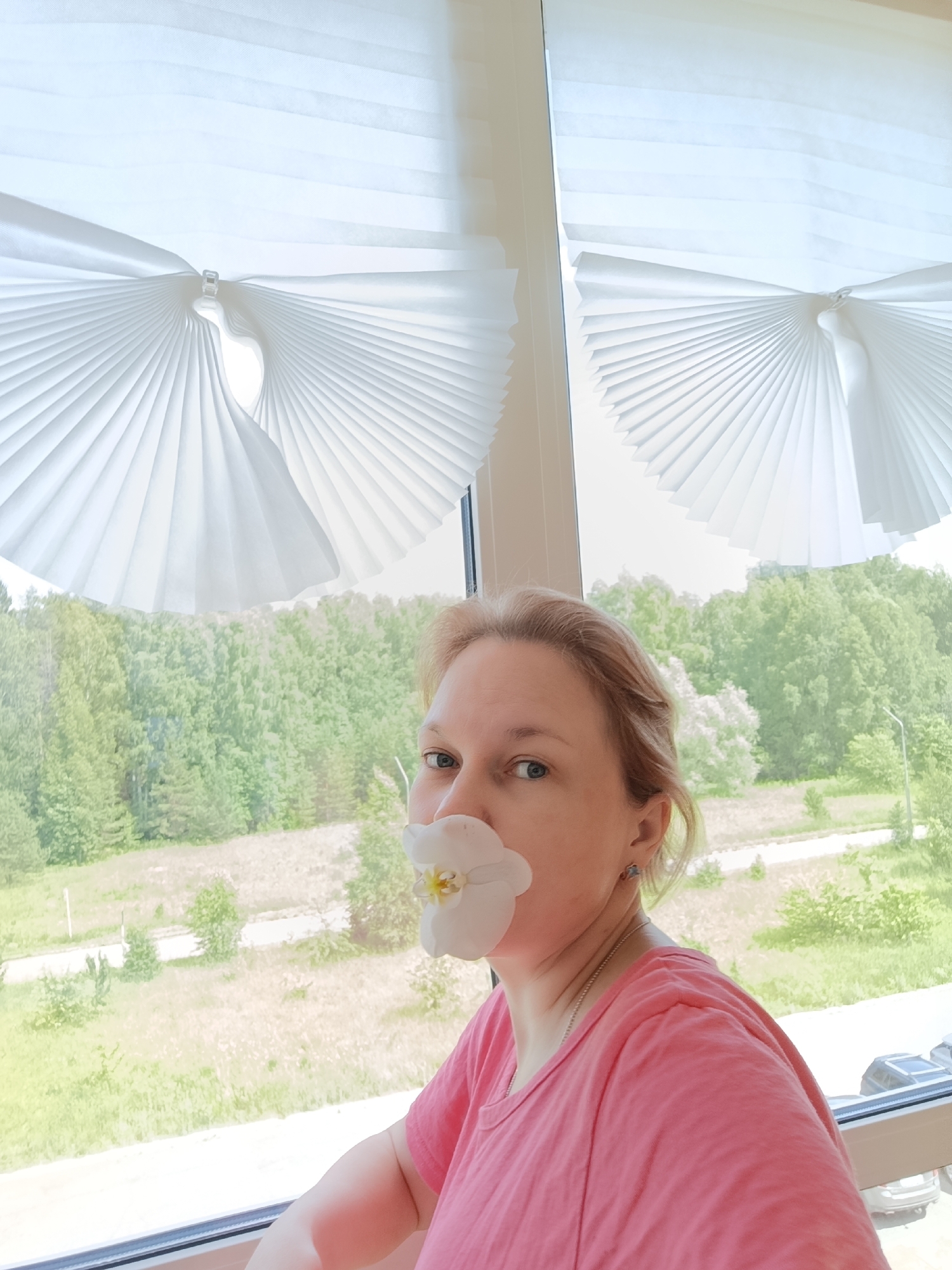 A Caucasian woman with blonde hair is positioned in the foreground, her face partially obscured by a white orchid petal held between her lips. She is wearing a pink t-shirt and a silver necklace. The woman is seated near a window, behind which a lush green forest and a grassy field are visible. Two white pleated blinds are installed on the window, their designs creating an aesthetic backdrop. The scene appears to be indoors, looking out onto a natural landscape. The image location is stated to be Марковский, Russia.