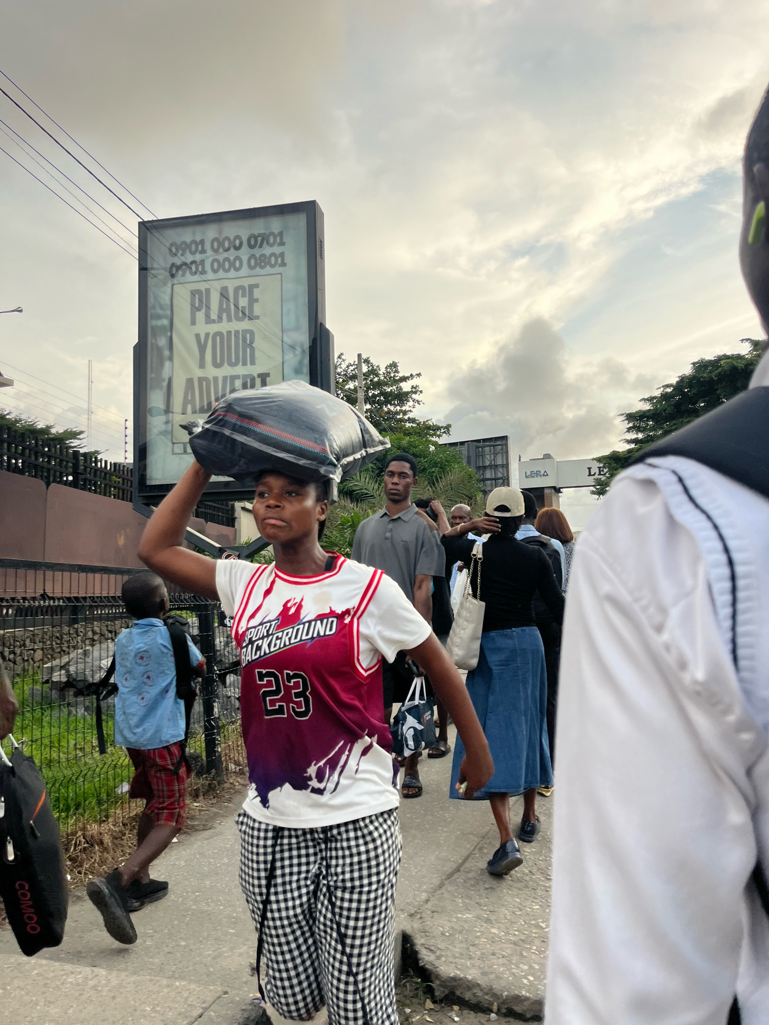A street scene in Lekki Phase I, Nigeria, shows a young woman walking towards the viewer, balancing a large black bundle on her head. She is wearing a white tank top with a red and purple graphic design and the number "23", along with black and white checkered trousers. Behind her, a young boy in a blue shirt and red and black plaid shorts walks away from the viewer, carrying a backpack. Several other people are visible in the background, including a man in a grey polo shirt and a woman in a black top and blue denim skirt. A large billboard with the text "PLACE YOUR ADVERT" and phone numbers is visible in the background. The environment appears to be a sidewalk next to a road with a metal fence and some greenery. The sky is overcast with clouds.