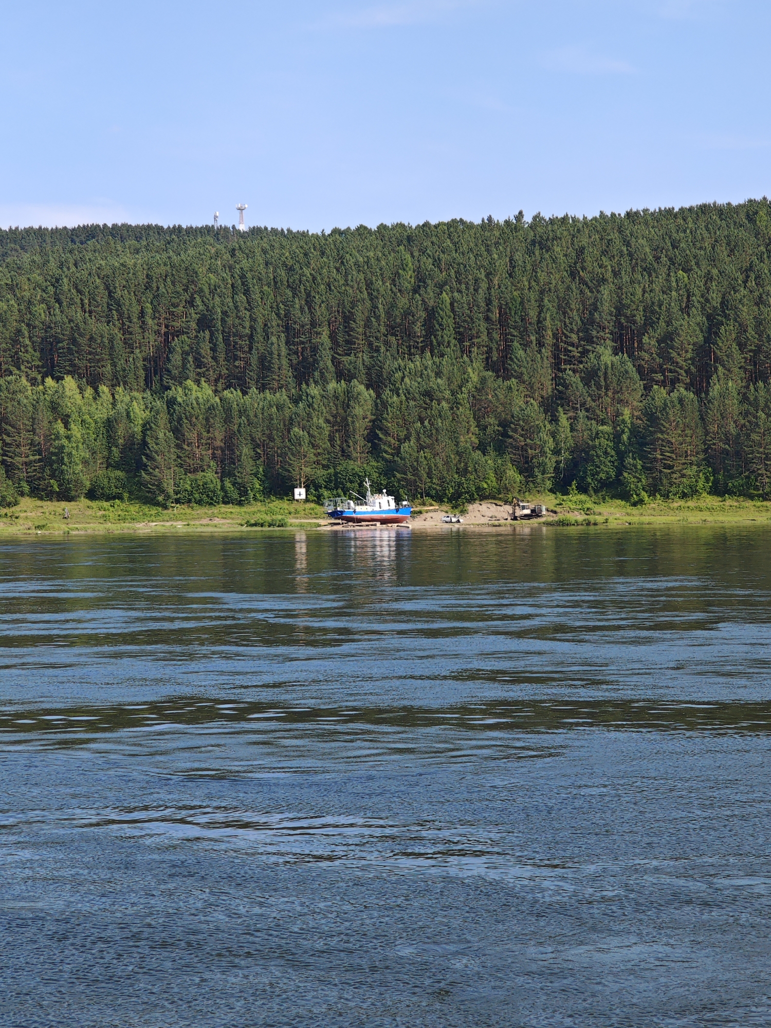 The image depicts a wide river in the foreground, with calm, rippling blue water reflecting the sky and surrounding greenery. On the far bank, a dense, green forest covers a sloping hillside. In the mid-ground, several communication towers are visible on top of the forested hill. Along the riverbank, at the base of the forest, a sandy shore is visible. A blue and white boat is partially on the shore, resting on some supports. To the right of the boat, there is a small excavator and a white car parked on the sandy bank. A sign with a white cross on a white background is also visible near the boat. The sky is clear and blue. The location is the Yenisei River near Predivinsk, Russia, a town situated on the right bank of the river.