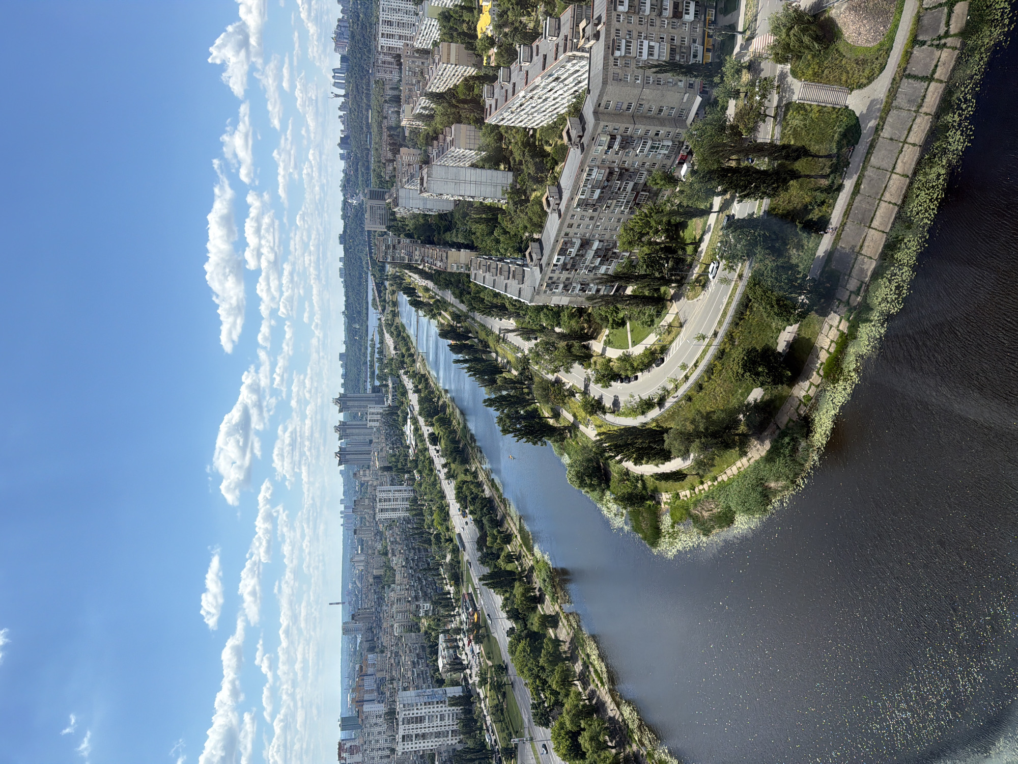 An aerial, high-angle view captures a wide, dark river flowing through a densely populated urban area in Kyiv, Ukraine, on a bright, cloudy day. The river occupies the left and lower portions of the frame, with its surface reflecting the light. Along the right bank of the river, a road curves, lined by a pedestrian walkway and trees. Beyond the walkway, a series of residential buildings, typical of Soviet-era architecture, are densely packed. These buildings are light-colored with many windows and balconies, interspersed with green trees and some smaller structures.
Further back from the river and buildings, a multi-lane highway is visible, with cars driving on it. In the distance, the cityscape extends with more residential buildings and some taller, modern high-rises. The horizon is marked by a hazy, green treeline and more urban structures. The sky above is a vibrant blue, dotted with fluffy white clouds. In the foreground of the river, aquatic plants are visible along the banks. A single cyclist is seen on the walkway near the river, and a small boat or kayak is on the water. The scene appears to be a residential district with significant infrastructure and a natural waterway.