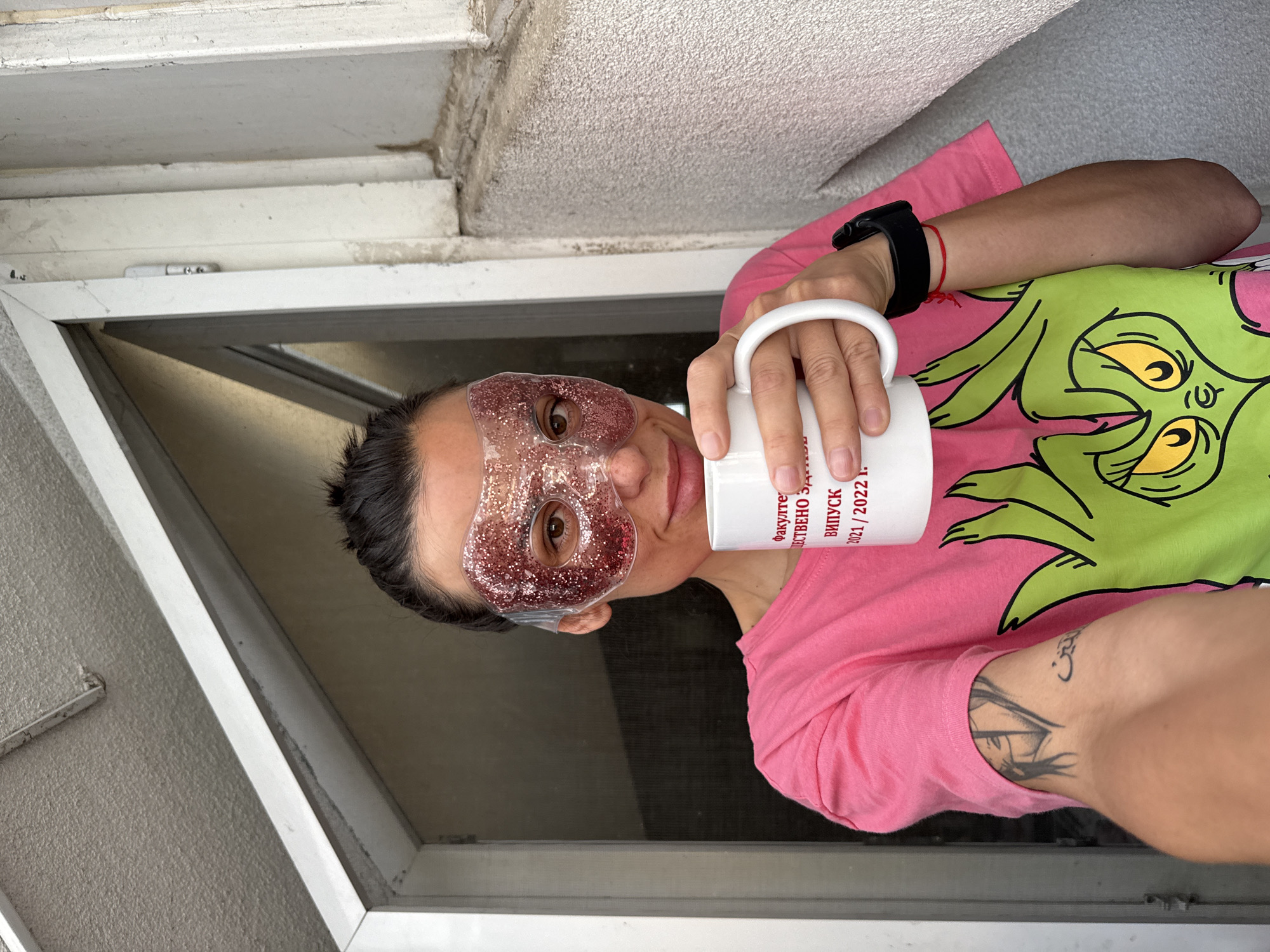 A person is taking a selfie indoors, possibly in Pleven, Bulgaria. The individual is wearing a glittery eye mask and holding a white mug with the words "факулте" and "ВИПУСК 2021/2022 г." printed on it. They are wearing a pink t-shirt with a green design, and a black watch is visible on their wrist. The person has a tattoo on their arm. They are positioned in front of a white-framed window. The wall and ceiling are off-white.