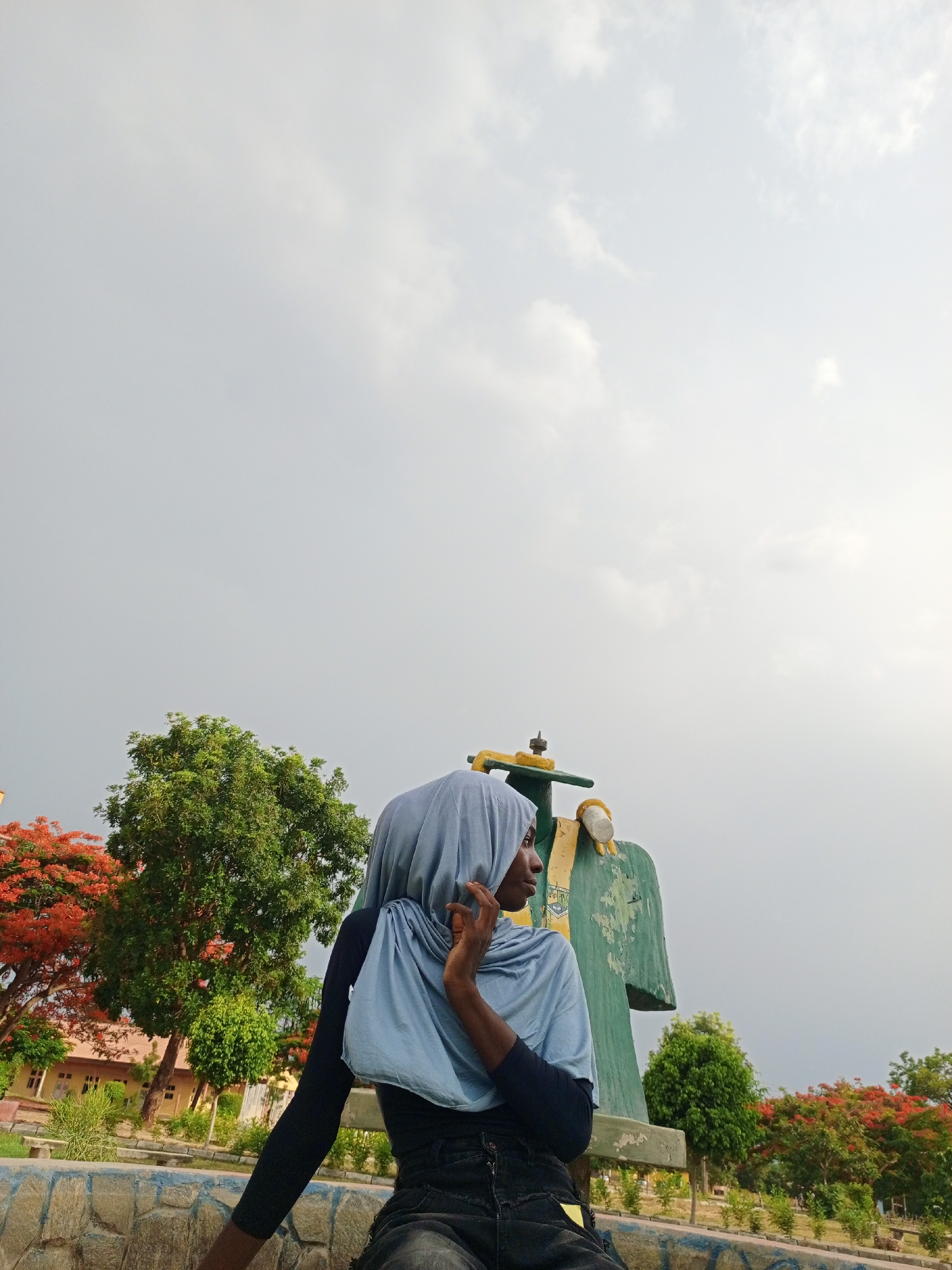 A woman wearing a light blue hijab is sitting on a bench outside in Unknown, Unknown. The sky is overcast. In the background, there are green trees with red flowers. There is a building with yellow walls behind the trees. Next to the woman is a large green sculpture with yellow accents.