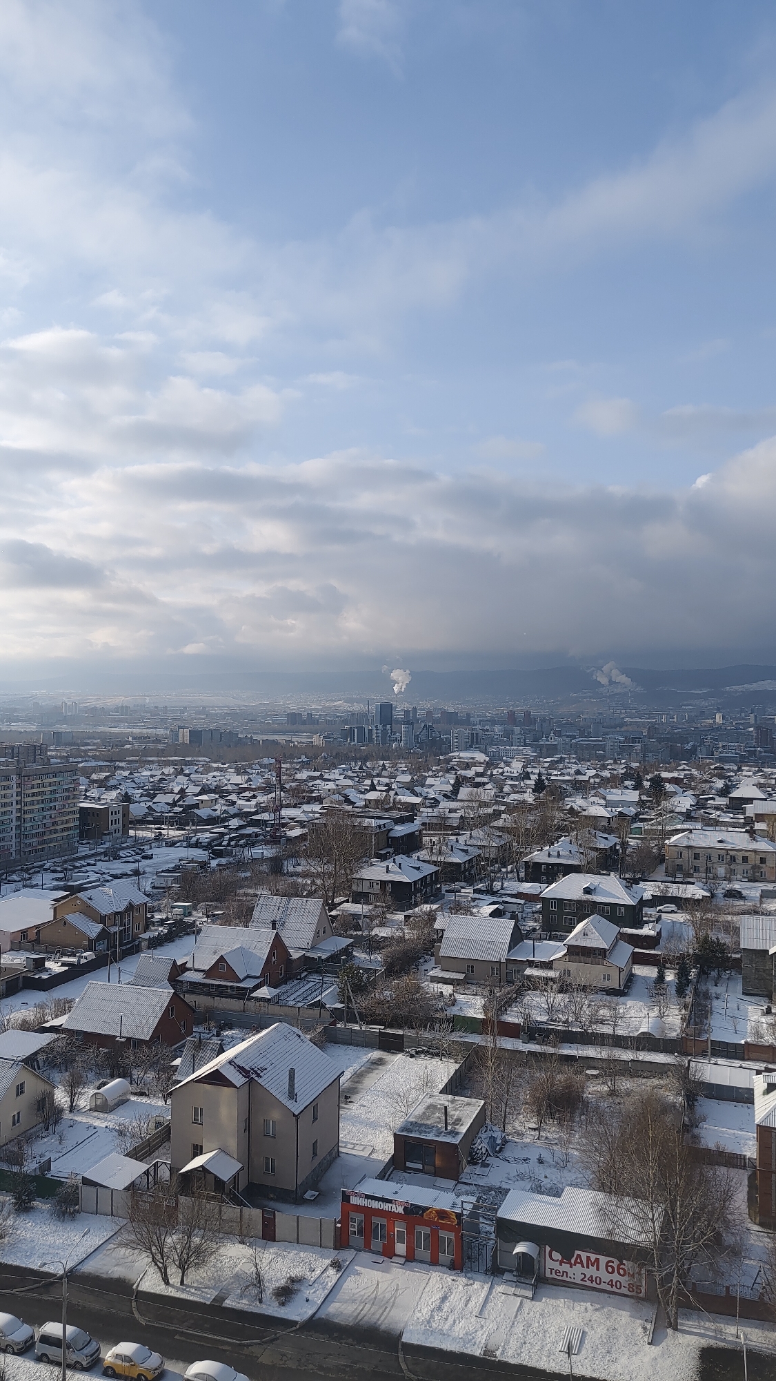 The image shows a panoramic view of Krasnoyarsk, Russia, in winter. The foreground is dominated by snow-covered residential buildings with peaked roofs. Streets are visible in front of the buildings, lined with bare trees. In the middle ground, there is a dense expanse of similar houses, blending into the cityscape. Further in the distance, a cluster of taller buildings forms a city center, partially obscured by haze or fog. Smoke plumes rise from industrial sites on the horizon. The sky is covered with a mix of blue patches and heavy, gray clouds. In the lower right corner, a storefront with red and orange facade is visible, with signage in Russian. The scene appears to be taken from an elevated position, possibly a high-rise building.