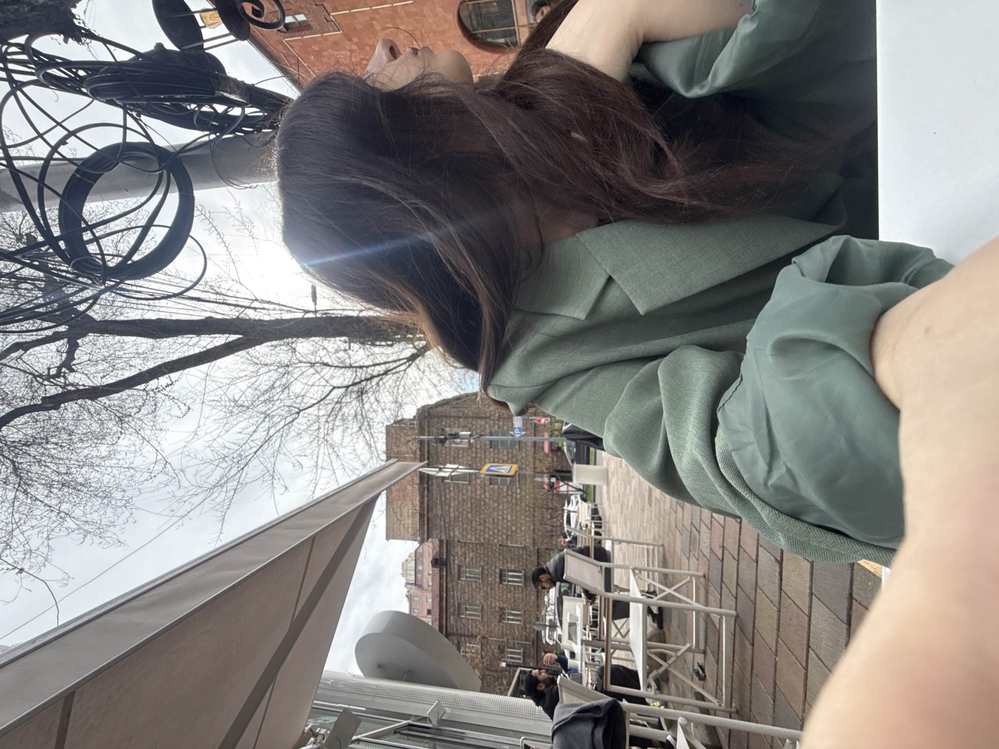 The image depicts an outdoor cafe scene in Yerevan, Armenia. The shot is taken from a low angle, looking up at a woman with long brown hair wearing a light green blazer. In the background, there is a multi-story building with stone facades and a pedestrian crosswalk sign visible. The cafe has white metal tables and chairs, with other patrons seated. There are bare trees and overhead wires visible, and the sky is overcast. A partial view of a white canopy covers some of the foreground.