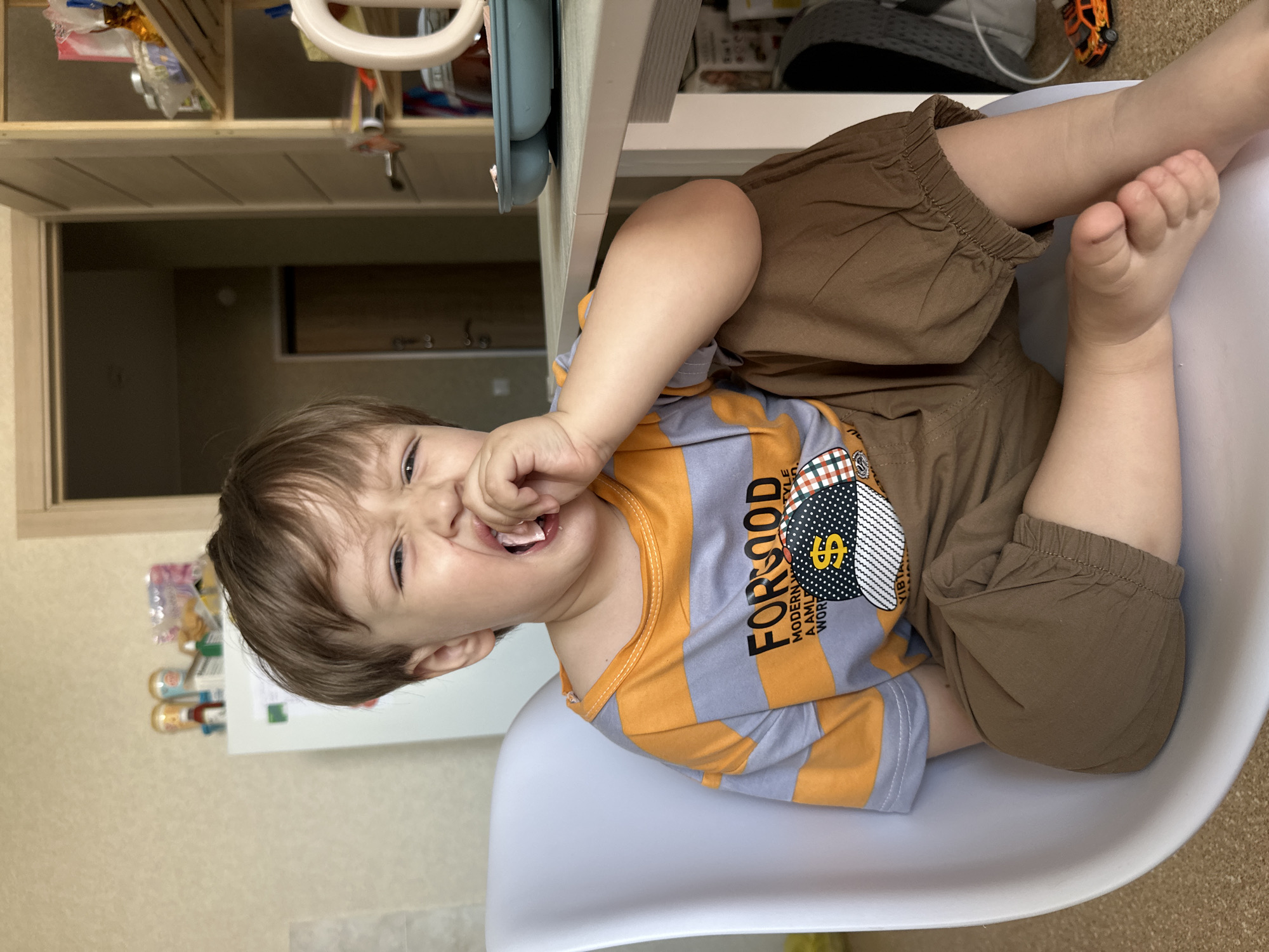 A young Caucasian boy, appearing to be between two and three years old, sits in a white plastic chair at a table. He has brown hair and is wearing a striped t-shirt with orange and grey bands, and brown pants. His right hand is in his mouth, and he is looking at the camera with his eyes squeezed shut and his mouth open. The background shows a kitchen with shelves holding various items, a refrigerator, and a doorway. A small toy car is visible on the floor near the table. The scene is indoors, in what appears to be a domestic setting. There is no visual information to specifically place this image in Ussurijsk, Russia.