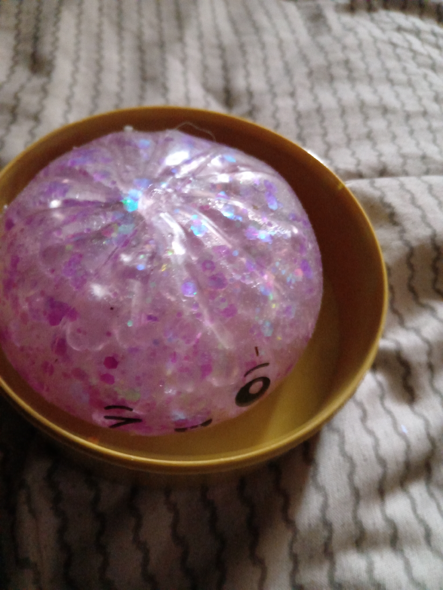A close-up, high-angle shot shows a light purple, glitter-filled squishy toy inside a yellow bowl. The toy has a slightly distorted appearance due to its material and the way it's contained. On the toy, there are crudely drawn black facial features: a smiling mouth and one visible eye. The bowl is placed on a surface covered with a patterned fabric, likely a bedsheet or tablecloth, with a wavy, striped design in light and dark gray. The lighting in the image is somewhat diffused, casting soft shadows. The scene appears to be indoors, in a residential setting. There is no indication of the city of Rudny, Kazakhstan, in the provided image or its crops.